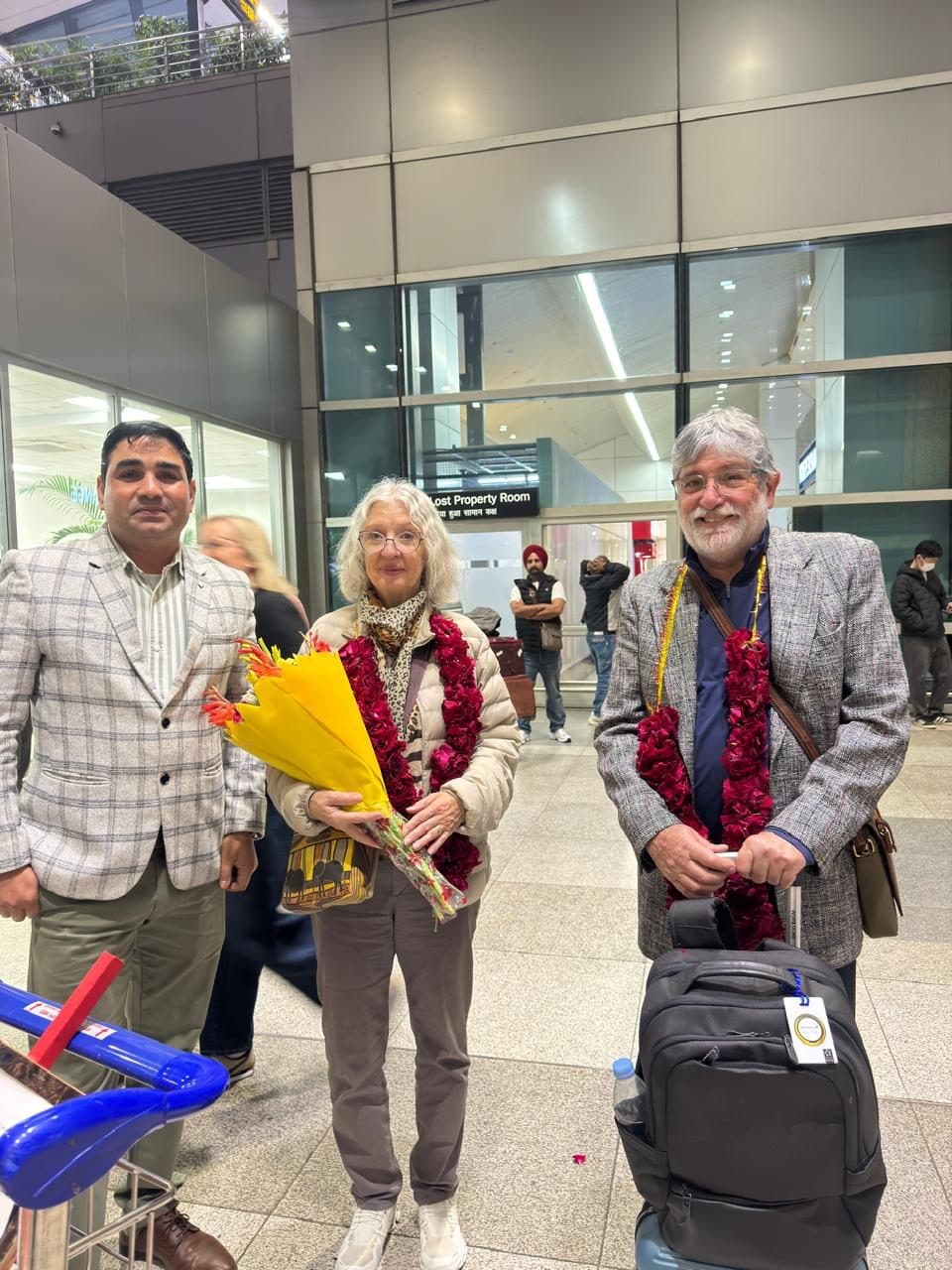 A guide presenting a bouquet to a woman guest while an older man beside her wears a garland, inside the arrivals hall