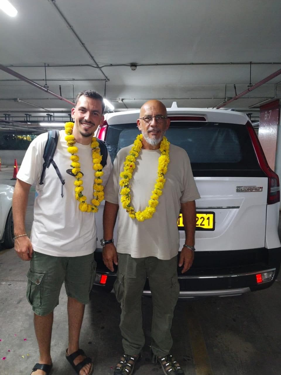 Two guests in yellow marigold garlands with their driver in the airport car park