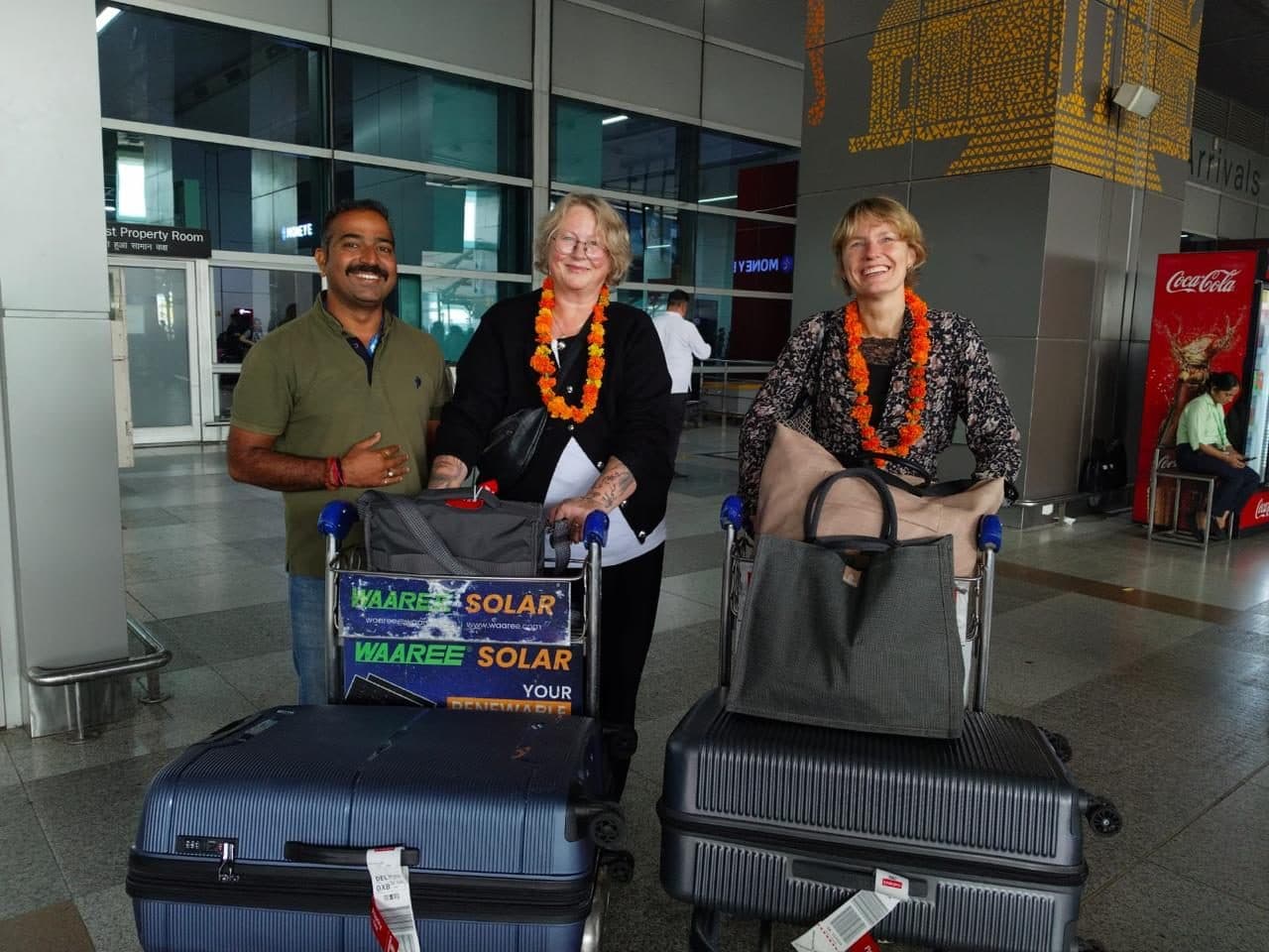 A guide with two women guests in garlands beside a loaded luggage trolley at Jaipur airport