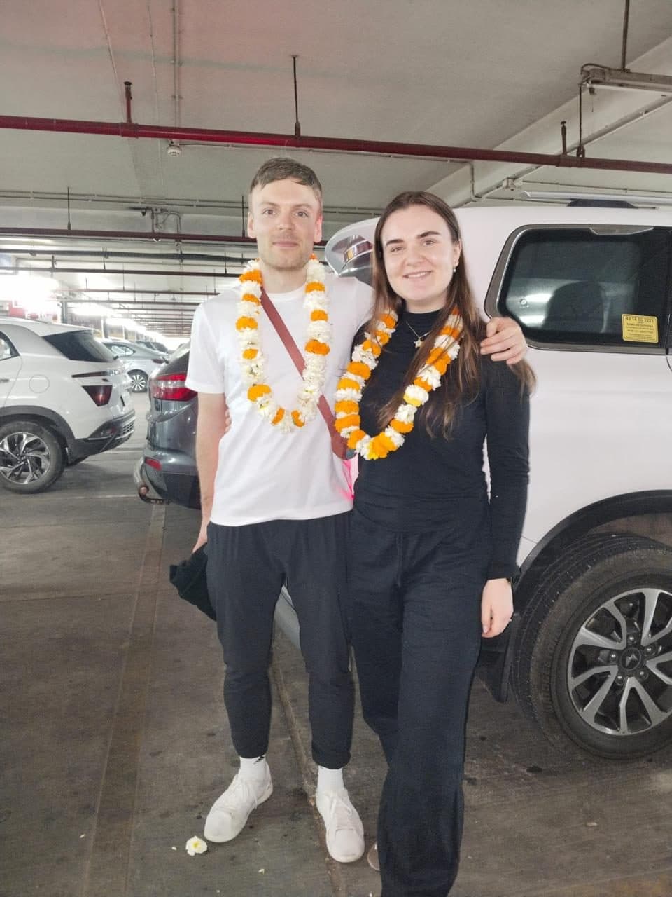 A guide with a male guest wearing a large marigold garland in the airport baggage area