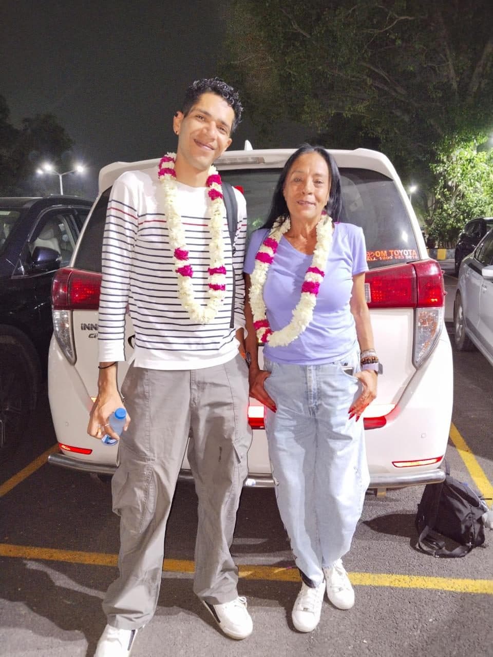 A young couple in garlands beside a white SUV at night, the boot open for their bags