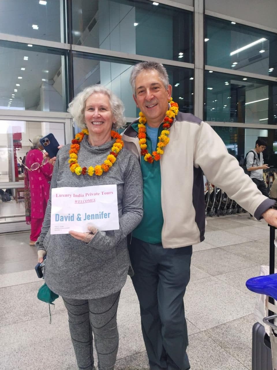 An older couple holding a name board reading David and Jennifer, with their guide at arrivals