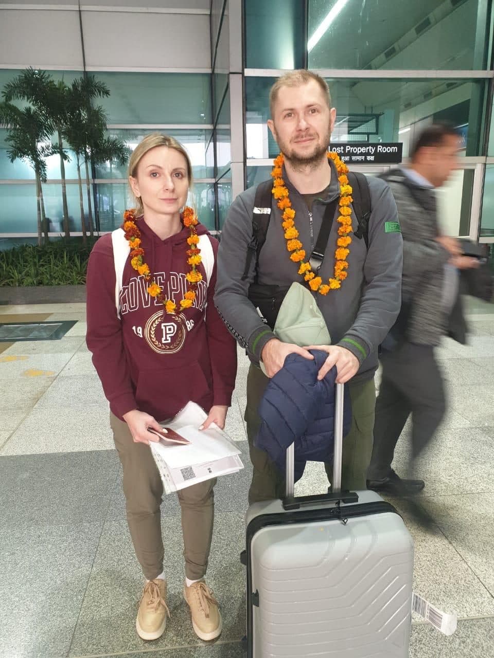 A guest and guide in garlands at the terminal, the guest's suitcase beside them