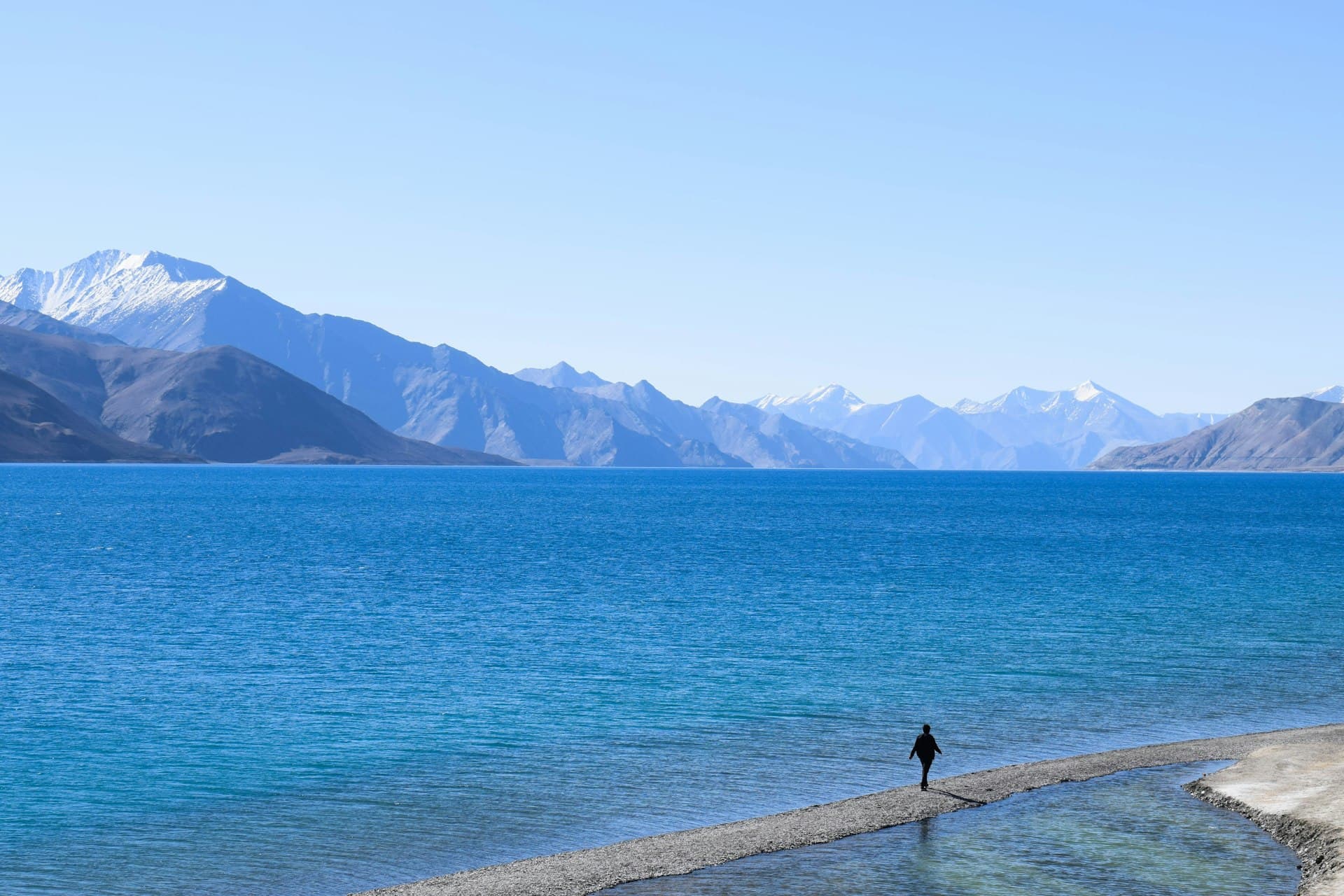 A person walking the sandy edge of Pangong Lake, the water shifting from pale green to deep blue toward the Ladakh mountains