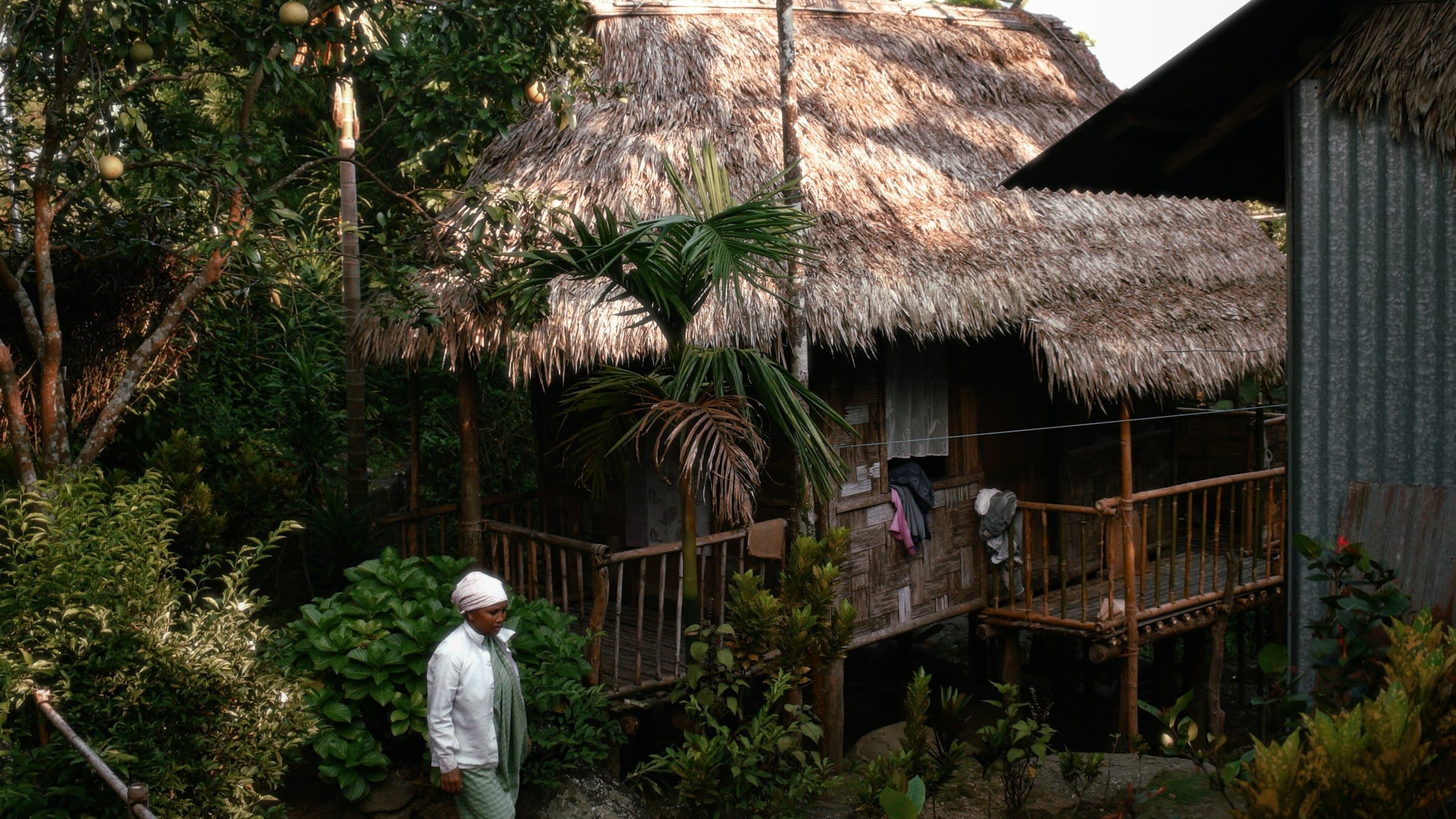A man stands beside a traditional thatched-roof Khasi home amid dense greenery in Mawlynnong village, Meghalaya.