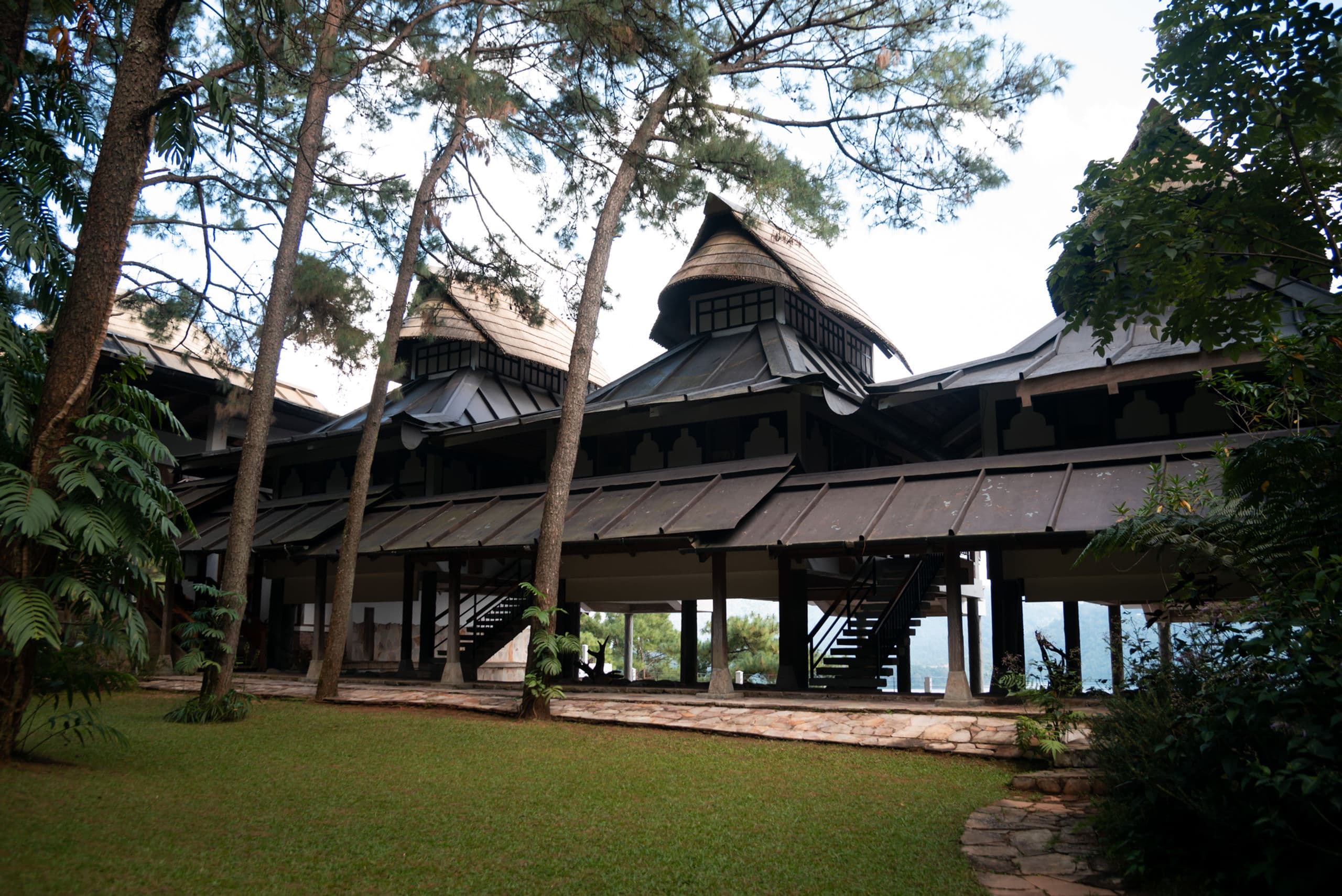 The long thatched Khasi-style main lodge building of Ri Kynjai seen across its lawn under tall pines.
