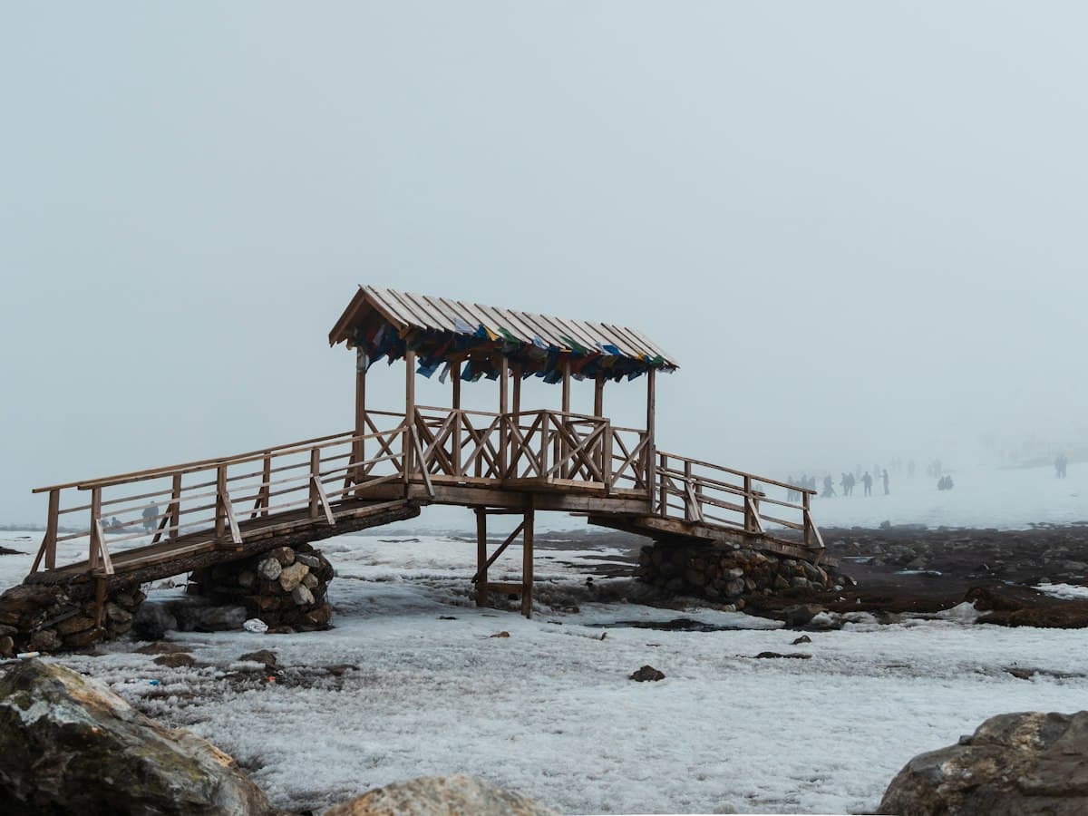 A rustic wooden bridge with a thatched roof over snowy ground amid barren mountains at Zero Point, North Sikkim.