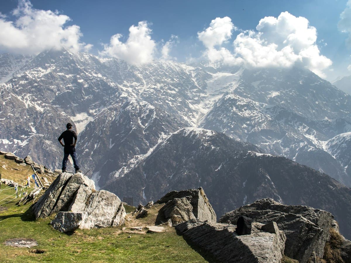 A hiker stands on a green mountain shoulder on the Triund trail with higher ridges ahead.