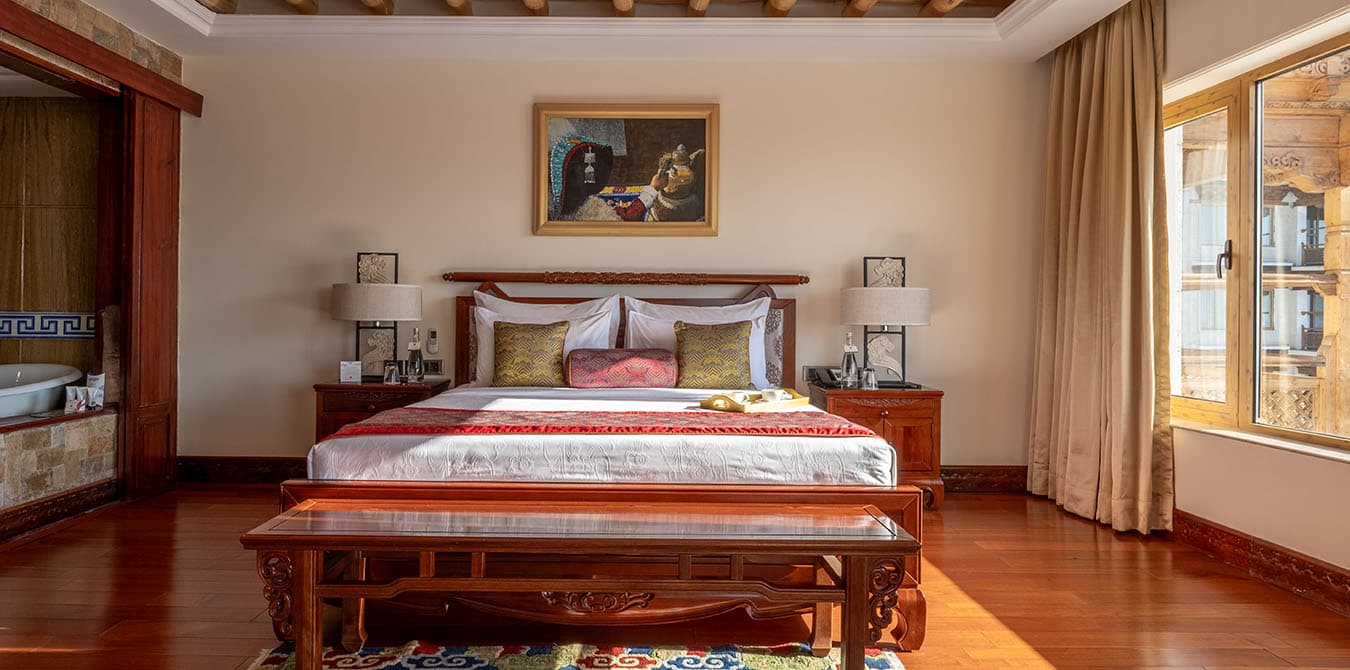Dragon Suite at The Grand Dragon Ladakh with a king bed below a framed painting, twin bedside lamps and a wooden bench at the foot