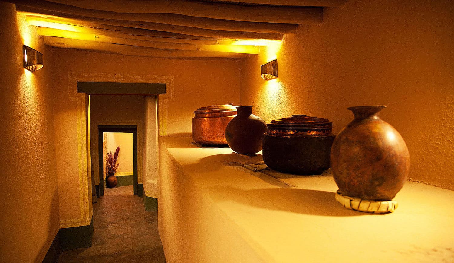 Museum passage inside Stok Palace lit by wall sconces, with antique copper and clay vessels displayed on a mud-plastered ledge