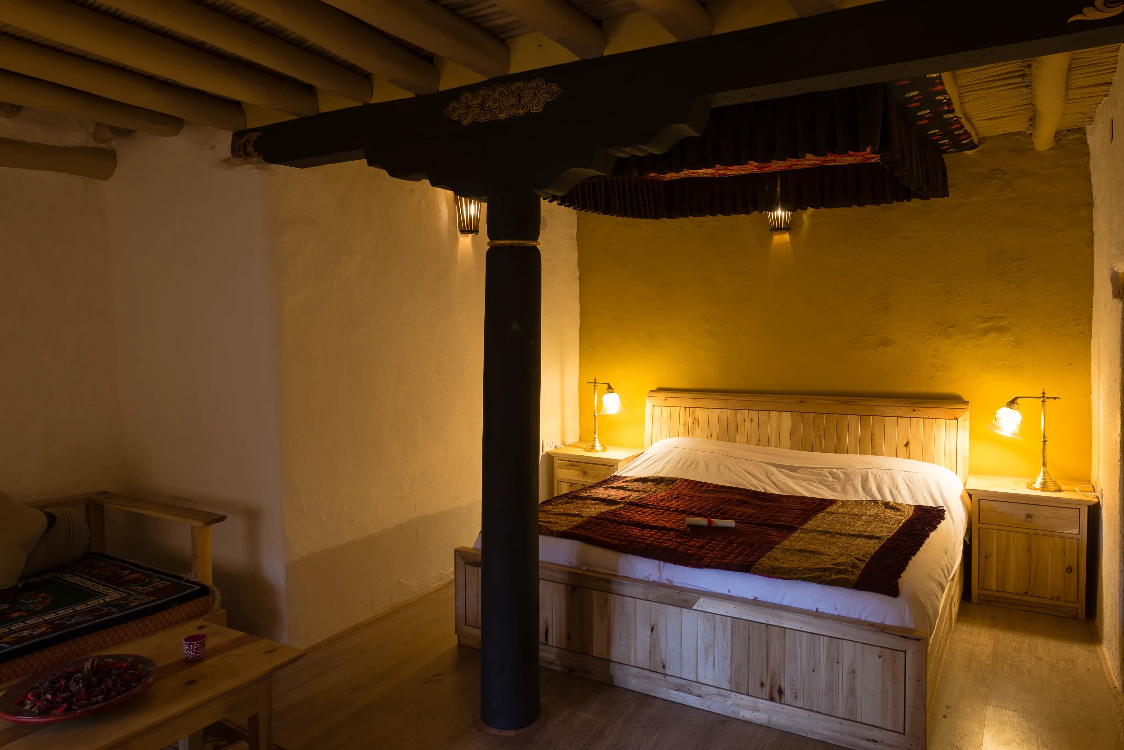 A suite at Stok Palace Heritage with a carved four-poster bed, whitewashed stone walls, exposed timber ceiling and a corner sitting area with bay windows