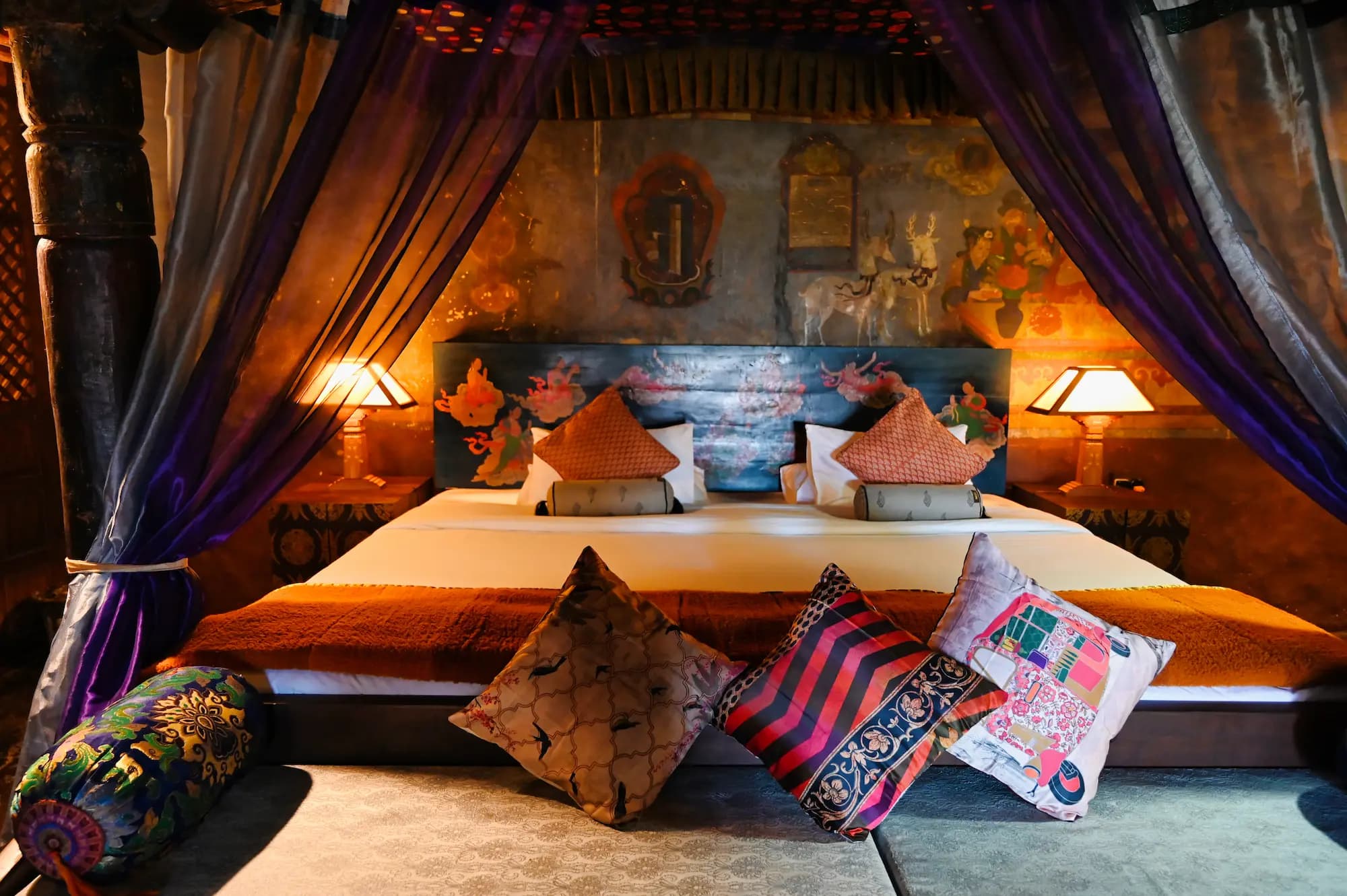 A royal suite at Stok Palace Heritage with a canopied bed dressed in Ladakhi cushions, hand-painted headboard wall and brass lamps