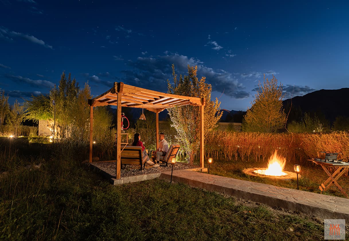 A wooden garden pergola with two lounge chairs among poplars and tall grasses at Lchang Nang Retreat, framed by Nubra Valley peaks