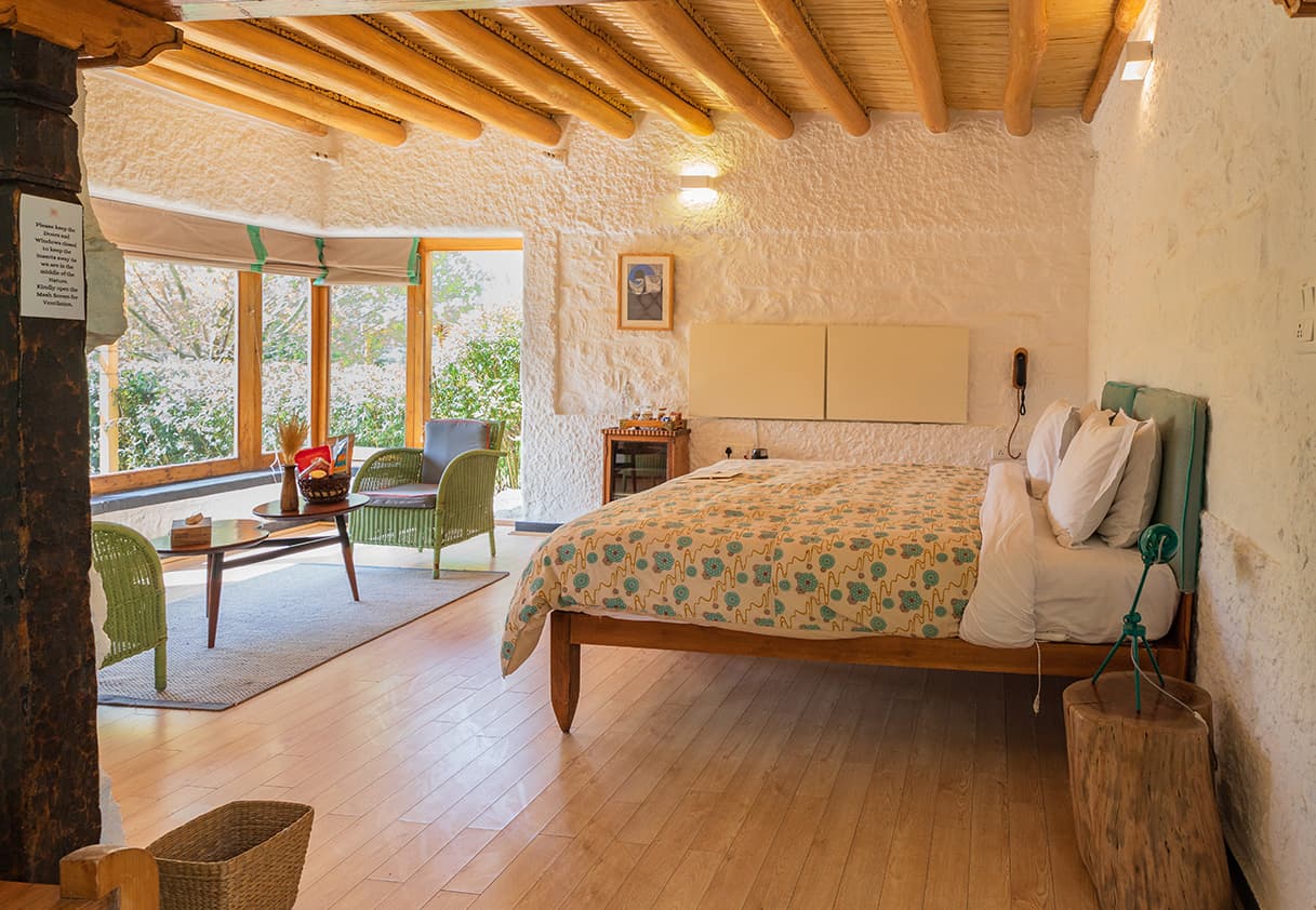 An eco-cottage bedroom at Lchang Nang Retreat with a low wooden bed, whitewashed stone walls, timber ceiling and a corner window seat facing the mountains