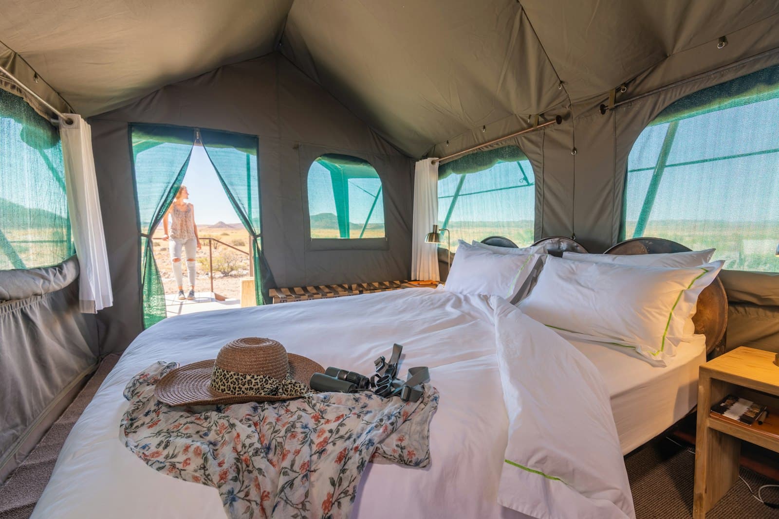 Interior of a luxury safari-style tent with a white-linen king bed, canvas walls and mesh windows opening to an arid mountain landscape