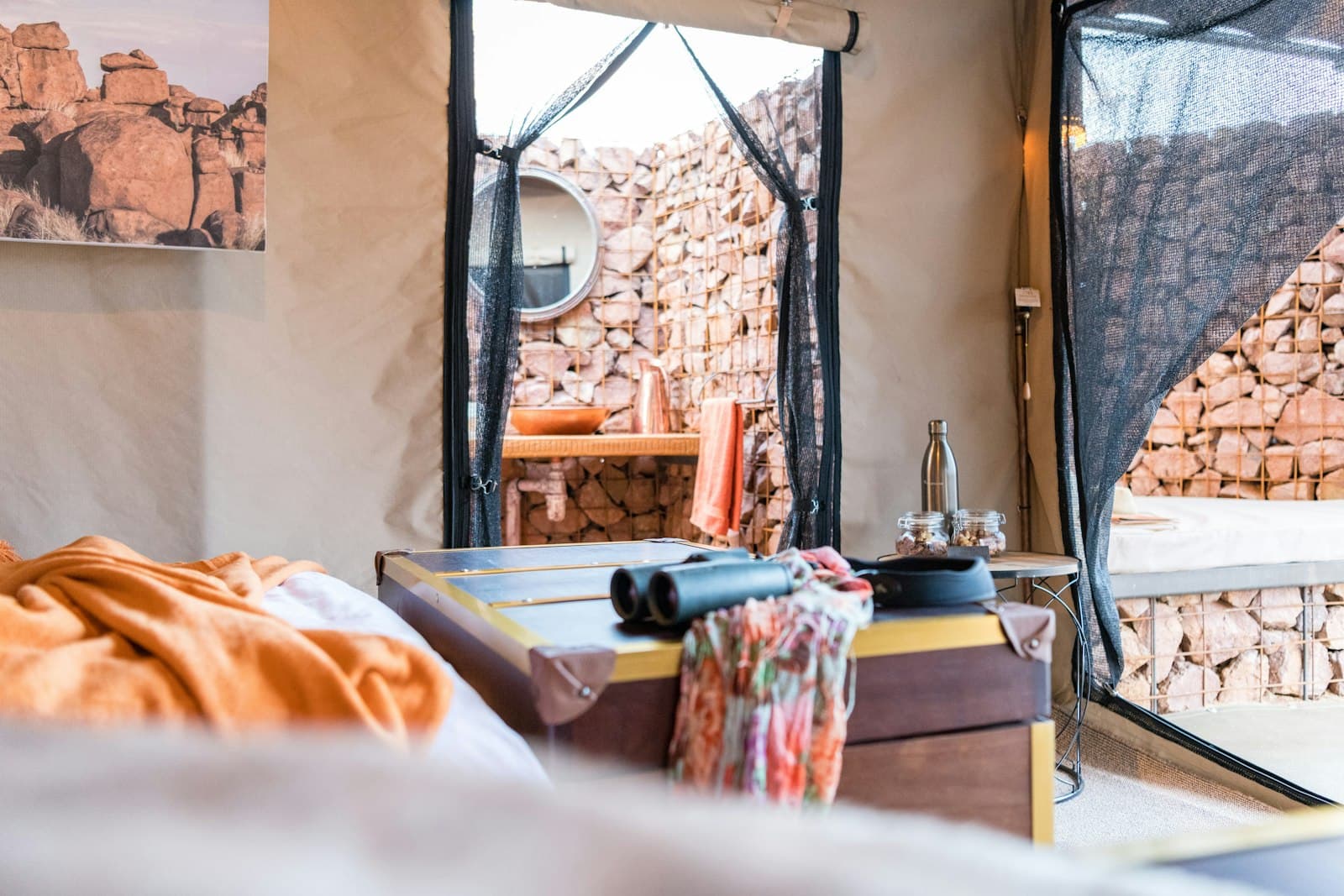 A tented bedroom with a made bed, a round mirror above a washstand and a canvas doorway framing a dry-stone wall outside