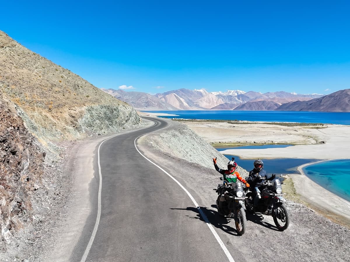 The winding mountain road drops toward the first blue sliver of Pangong Tso, riders pausing where the lake opens up between the ridges.