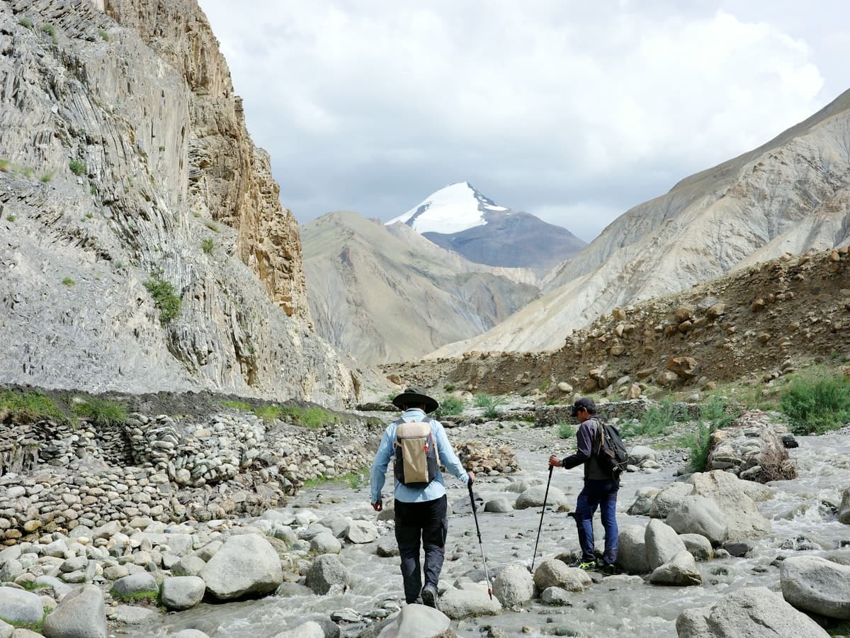 Two trekkers cross a rocky mountain stream beneath Kang Yatse on the Markha Valley trail in Ladakh.