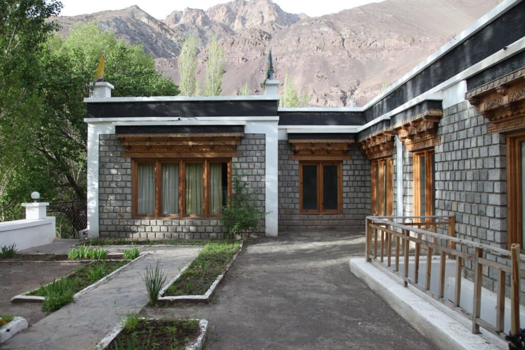 Exterior of The Apricot Tree hotel showing its wood-panelled Ladakhi facade and a garden path, with mountains behind