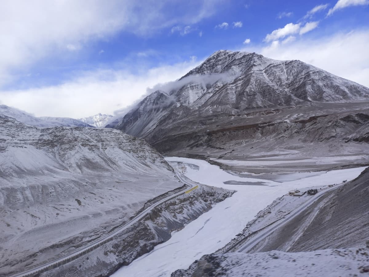 A river winds beneath a broad snow-capped massif, echoing the Suru Valley opening toward the twin peaks of Nun and Kun.