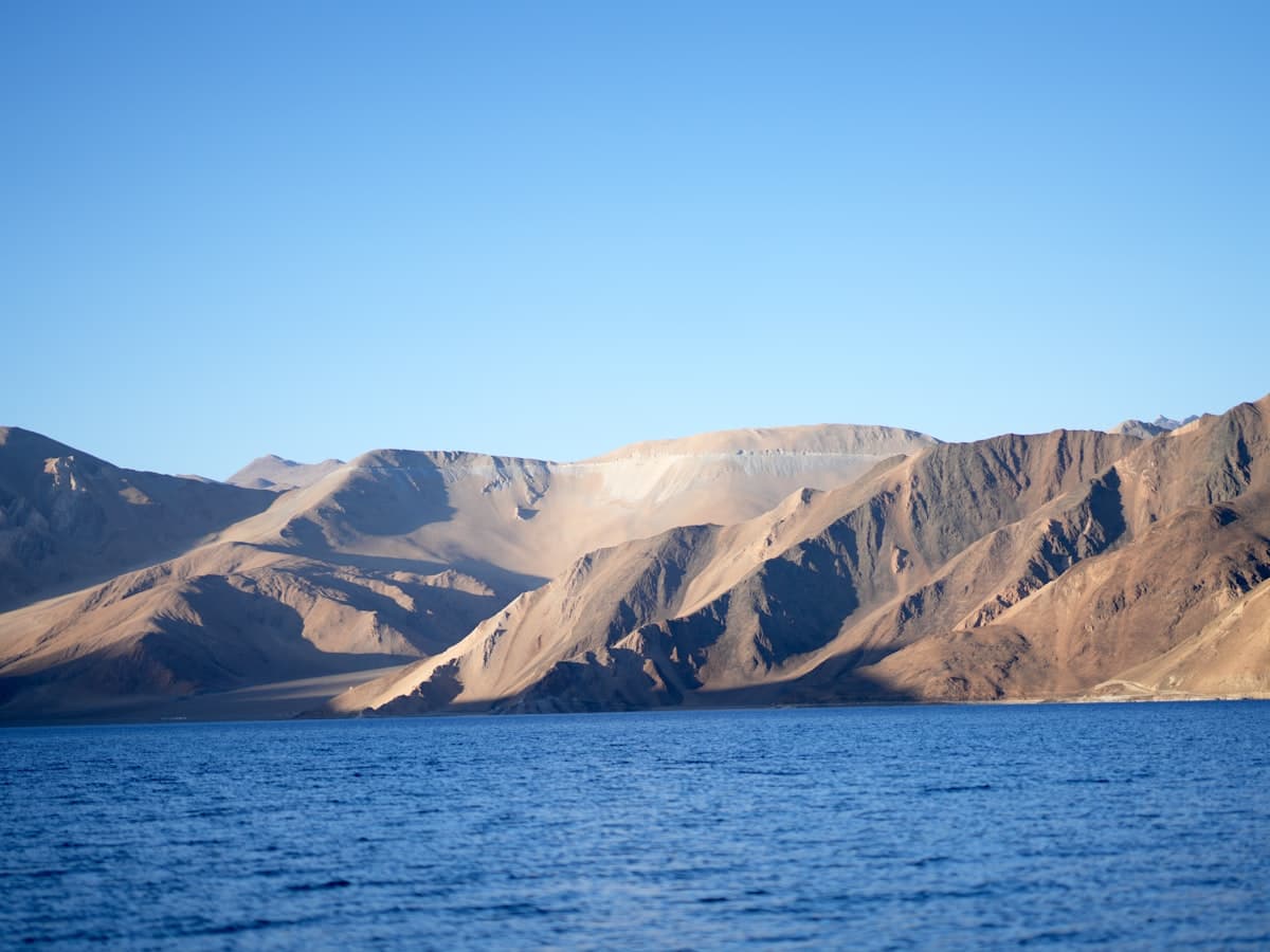 A vast still blue lake laps a barren shoreline below sunlit treeless mountains at high altitude.