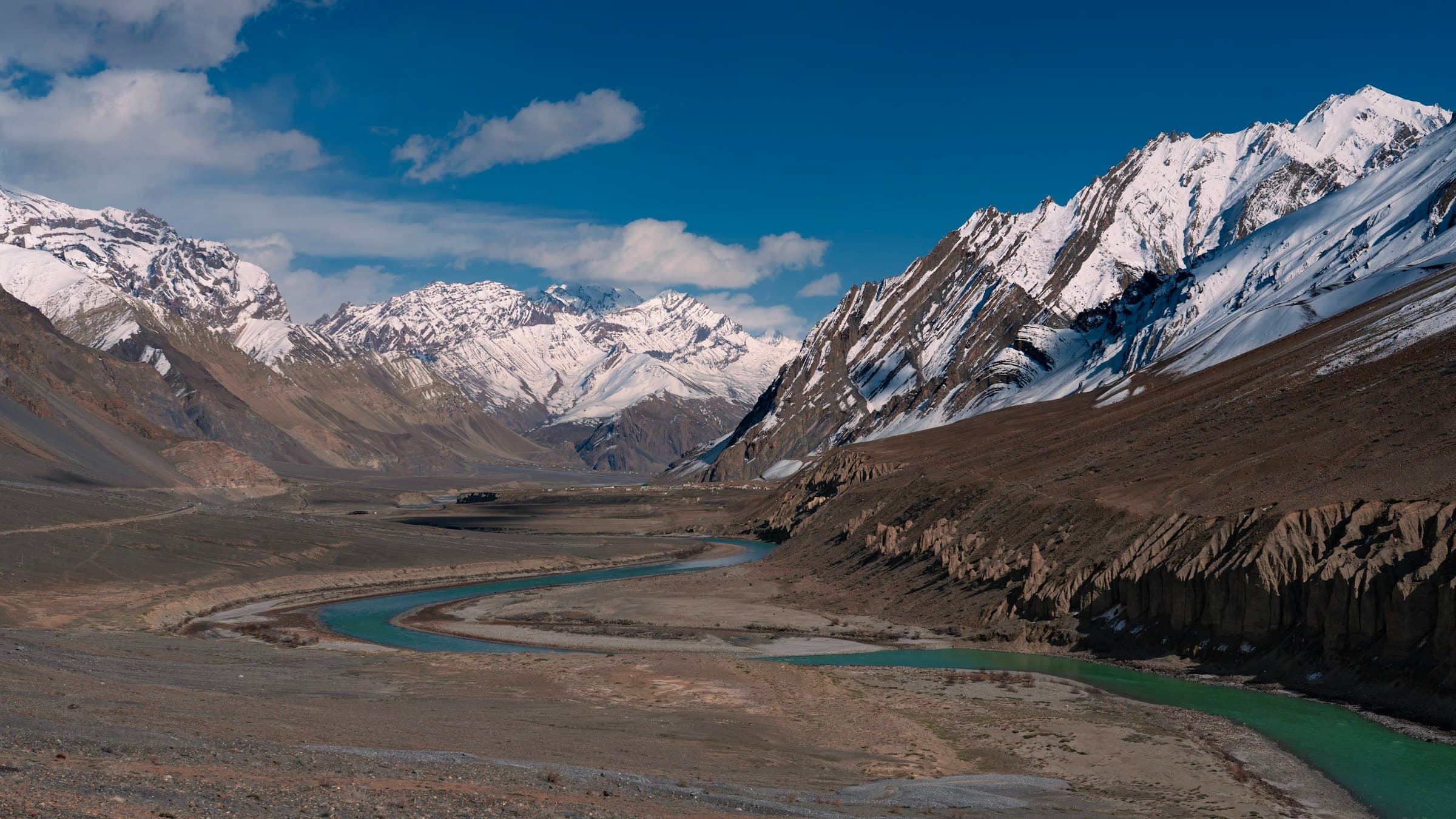 A river snakes through a broad green valley beneath a line of snow-capped Himalayan peaks in western Ladakh.