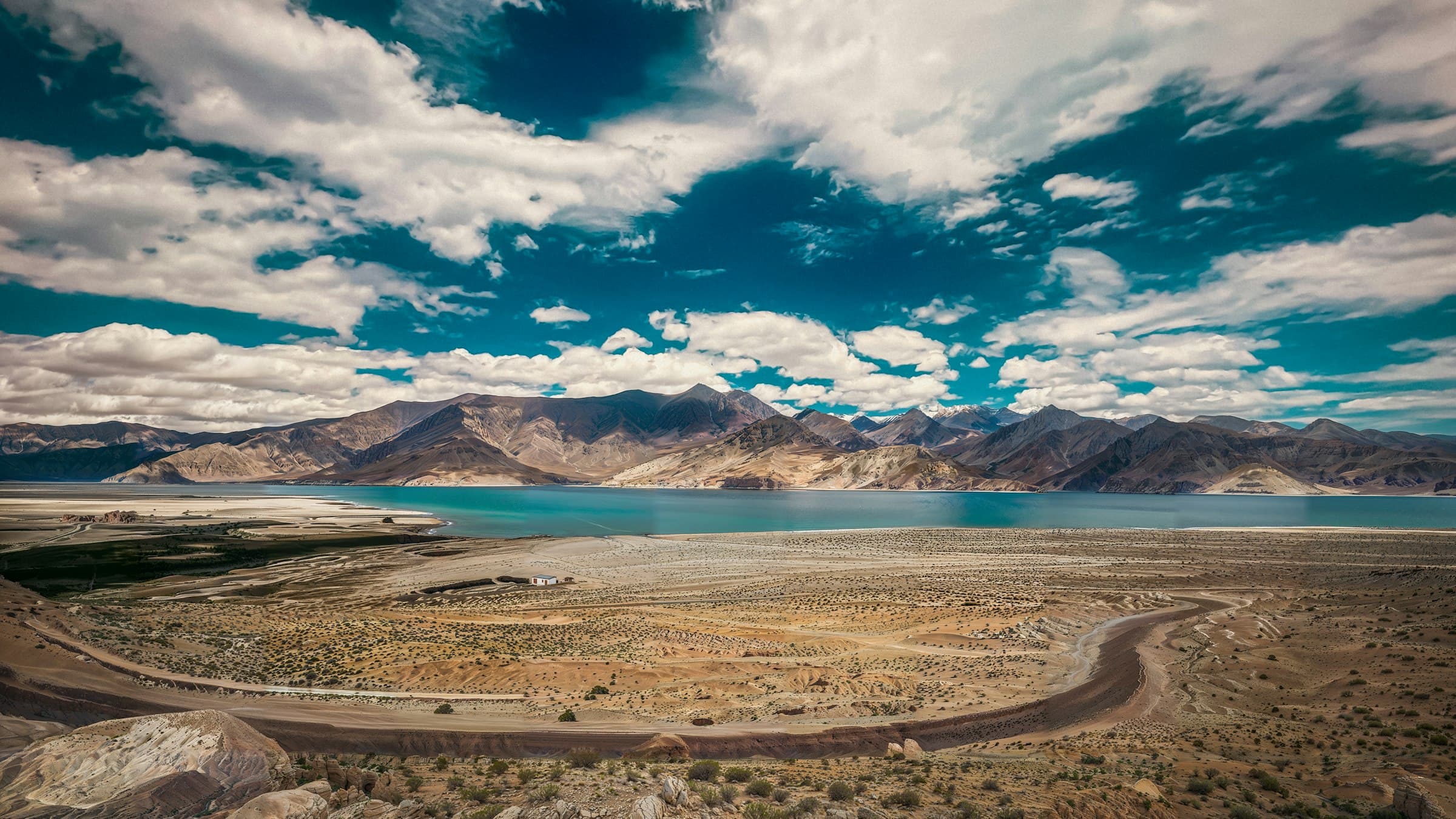 A turquoise lake and a curving desert road sit framed by rugged bare mountains on the Changthang plateau.