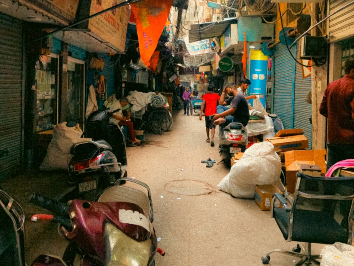 A crowded, narrow lane in Old Delhi hemmed by shopfronts, with scooters and pedestrians threading past under a canopy of tangled overhead wires.