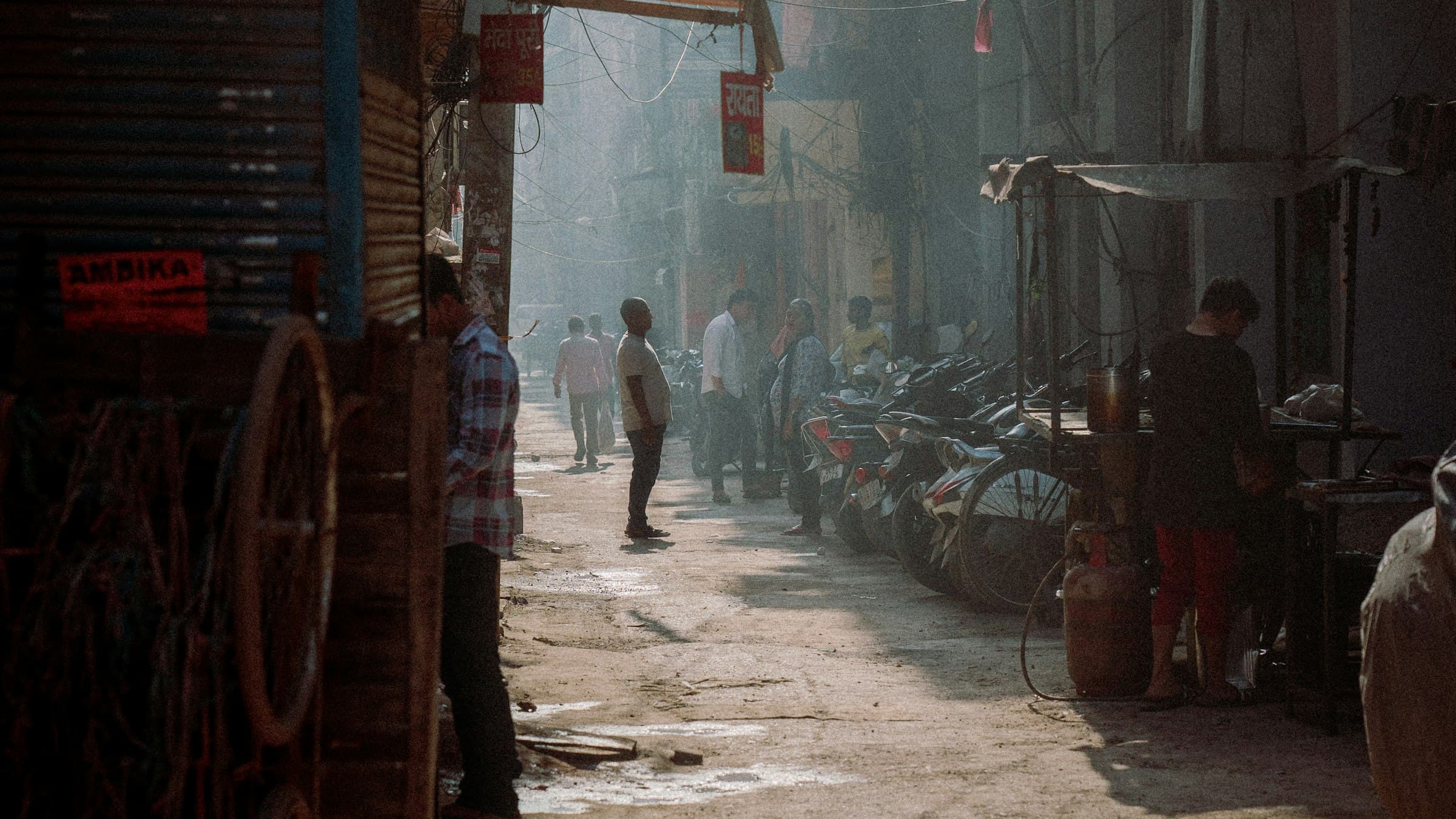 A man walking a narrow Old Delhi street below dense, close-packed multi-storey buildings and their web of overhead cables.