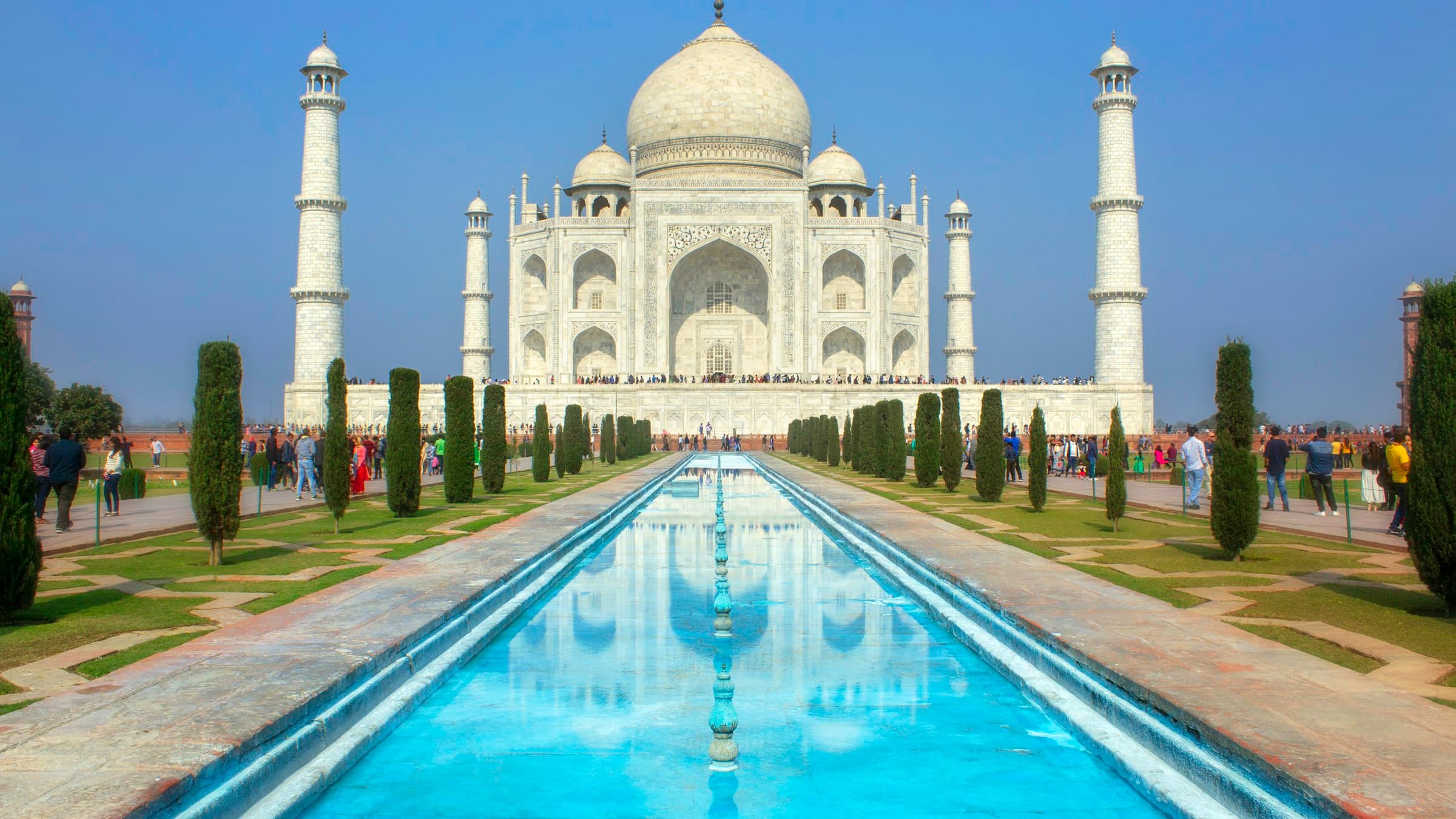 The Taj Mahal mirrored in the long rectangular reflecting pool of its formal garden, cypress trees flanking the walkway.