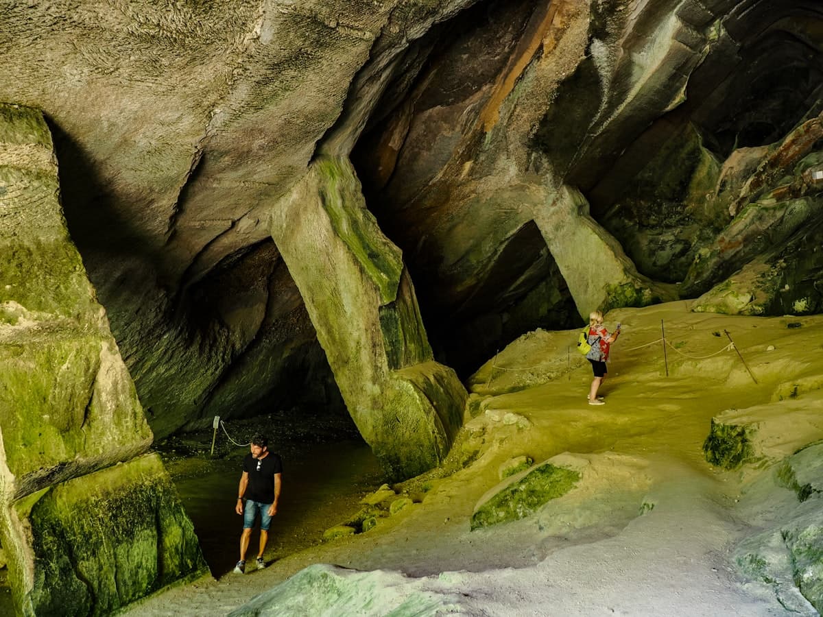 Two people exploring a large cave with mossy rock formations underground.