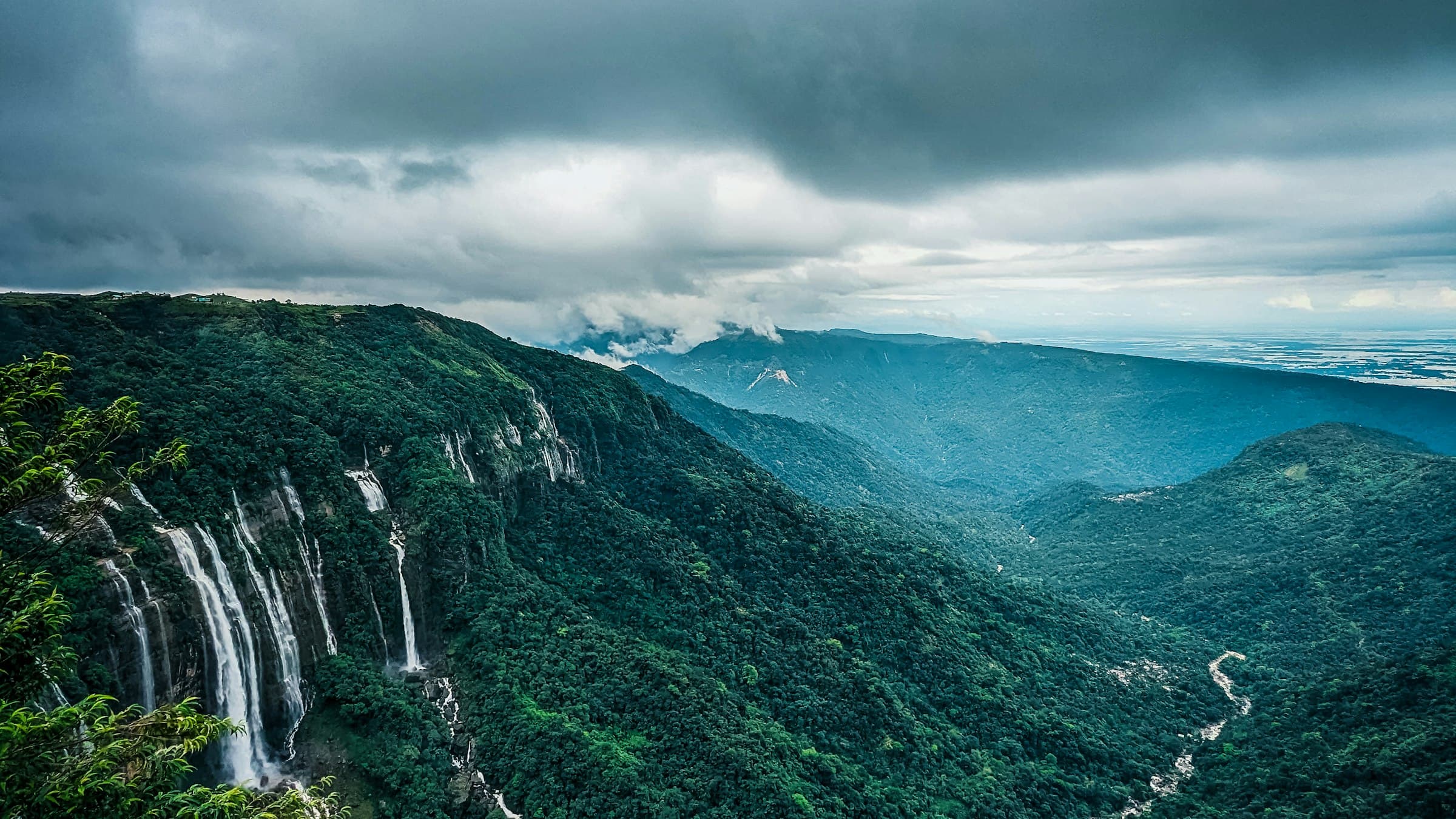 A broad green Cherrapunji valley with a waterfall threading down a far cliff.