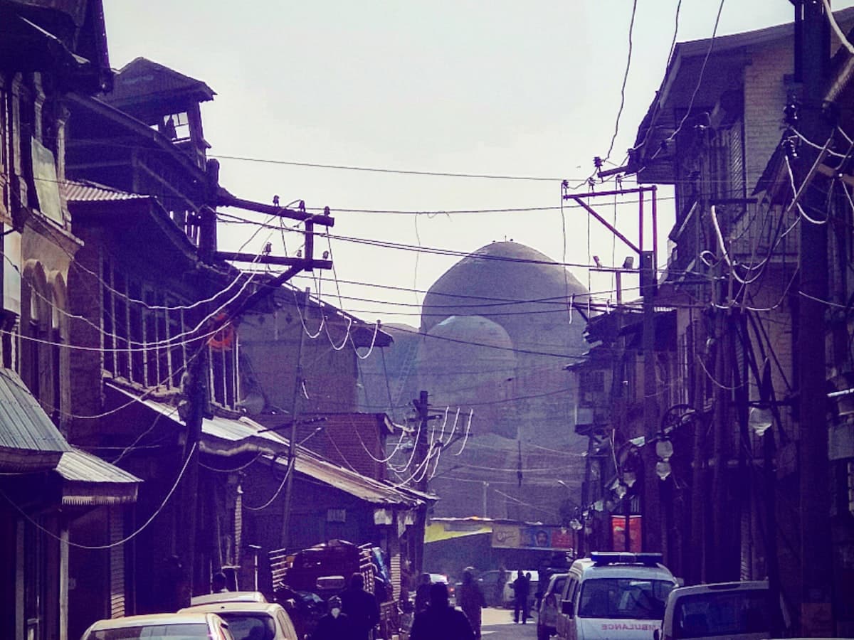 A man walks a street in Srinagar's old city past parked cars and low buildings.