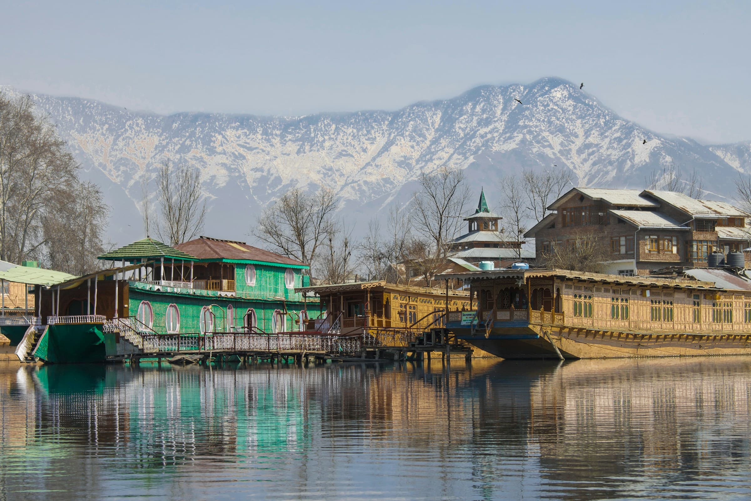 A green-and-brown wooden Kashmiri houseboat on Dal Lake below a snow-covered mountain.