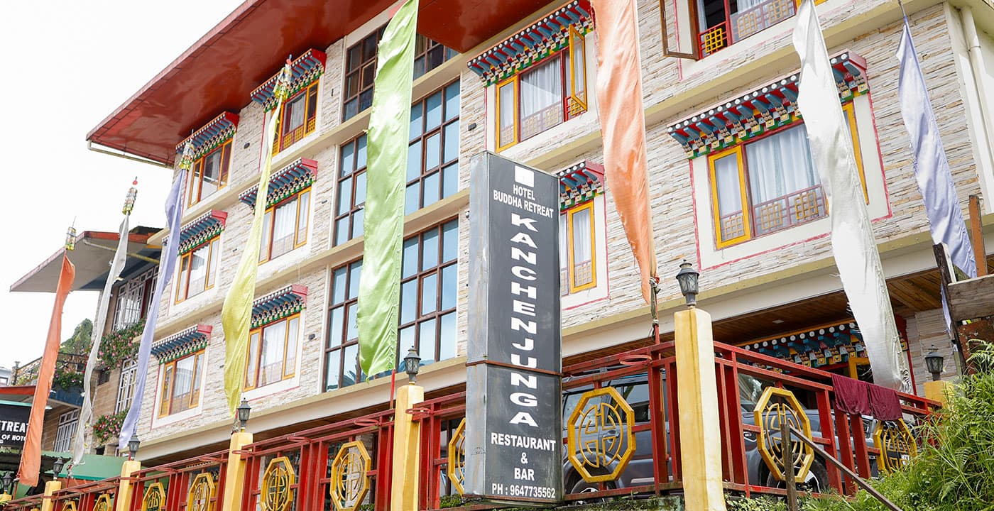 The Bhutanese-trimmed stone facade of Hotel Buddha Retreat in Ravangla, with prayer flags and its Kanchenjunga Restaurant & Bar signage.