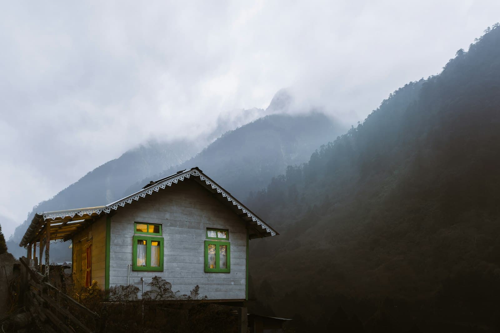 A traditional Sikkimese timber village house with carved bargeboards and green window frames below mist-wrapped Himalayan peaks, near Lachen.