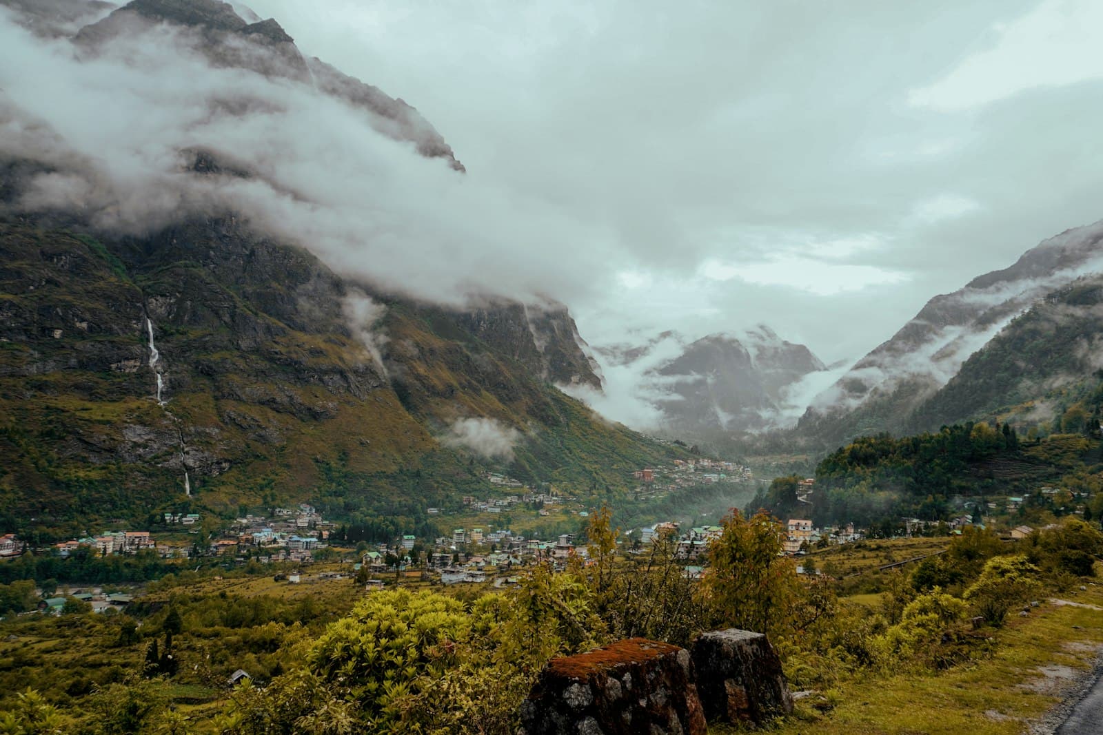 A North Sikkim mountain town spread across a green valley floor with a waterfall and cloud-wreathed Himalayan ridges above.