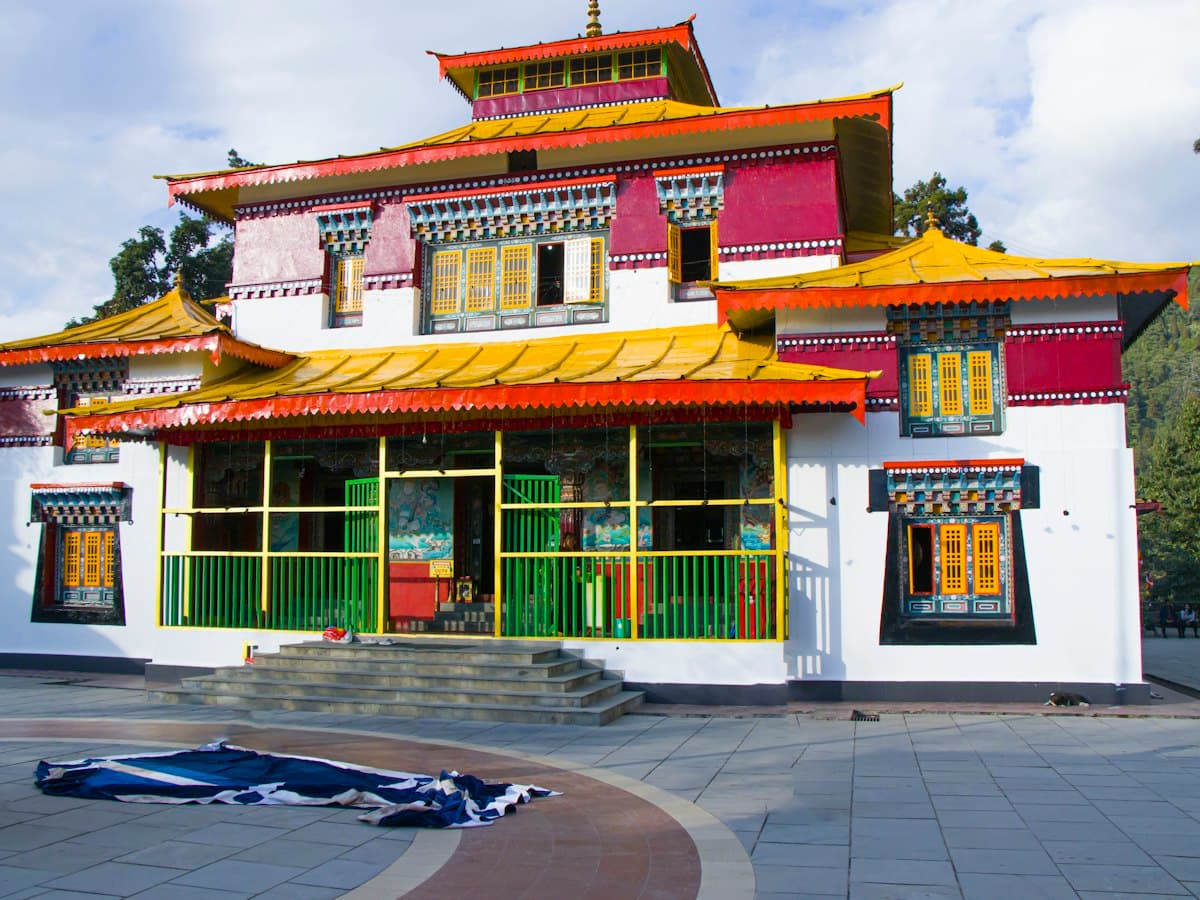 A white monastery building with yellow and red roofs on a forested ridge near Pelling, West Sikkim.