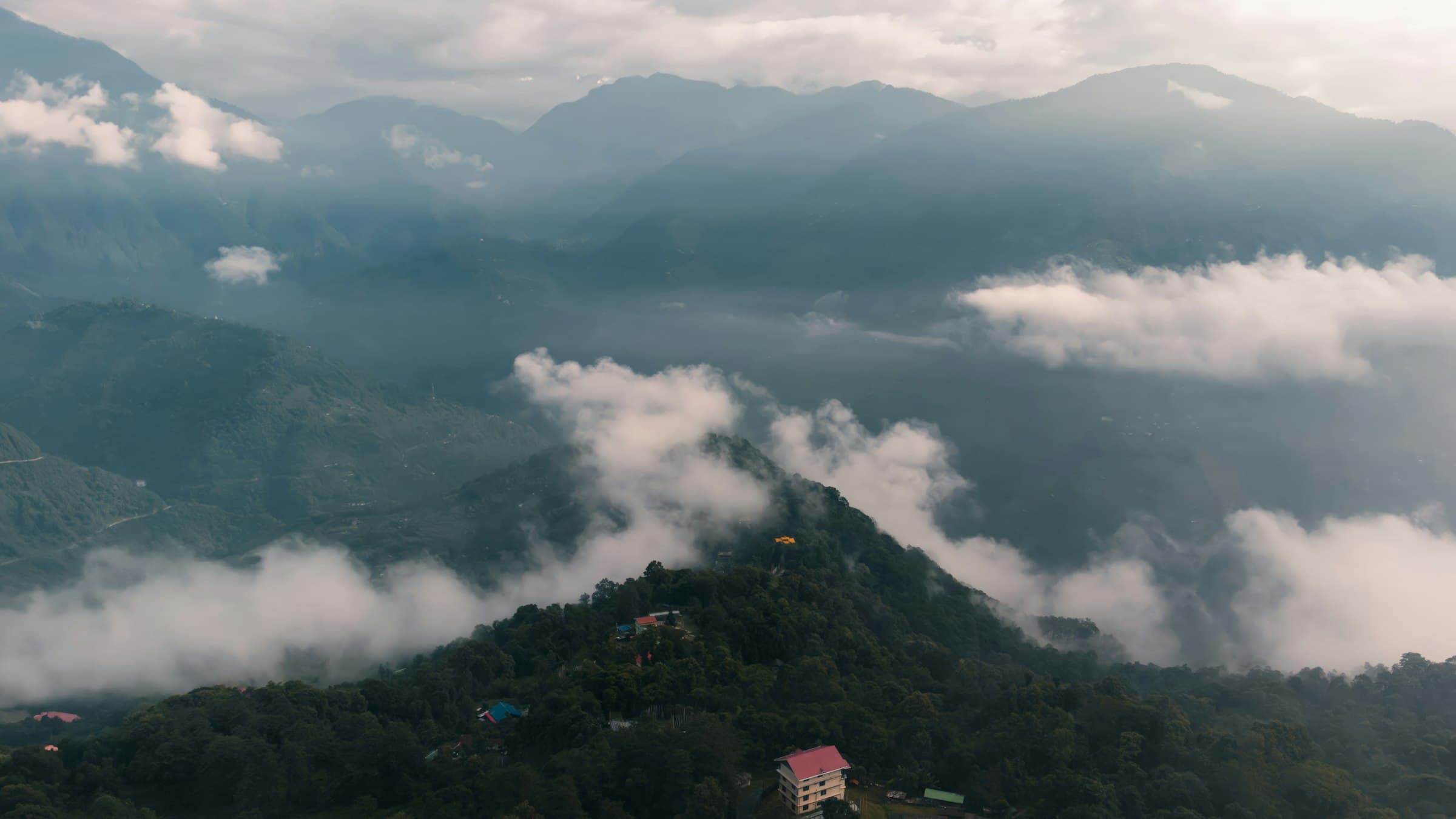 An aerial sweep over the forested ridges and scattered settlements around Pelling, West Sikkim, with cloud below the peaks.