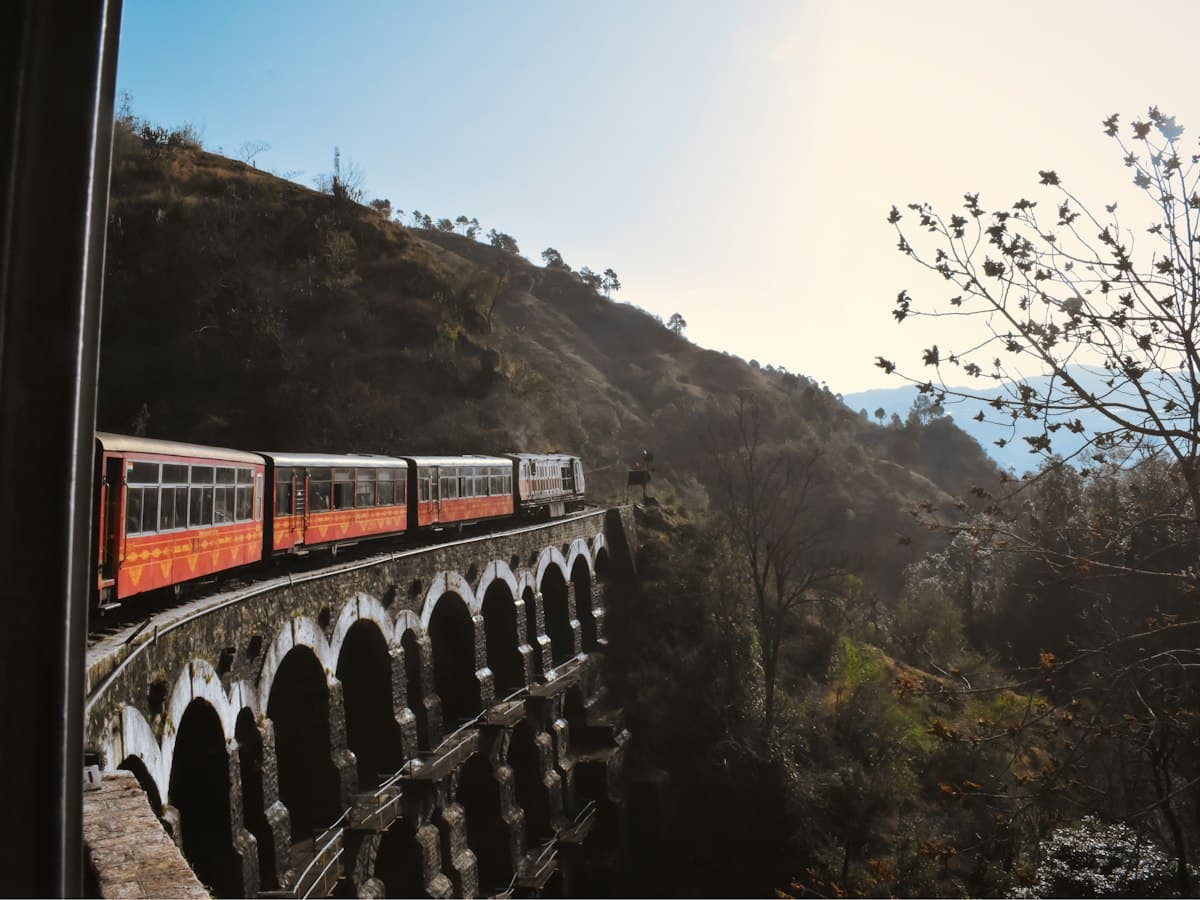 A narrow-gauge train crosses a stone arch viaduct on the Kalka-Shimla railway through pine-covered hills.