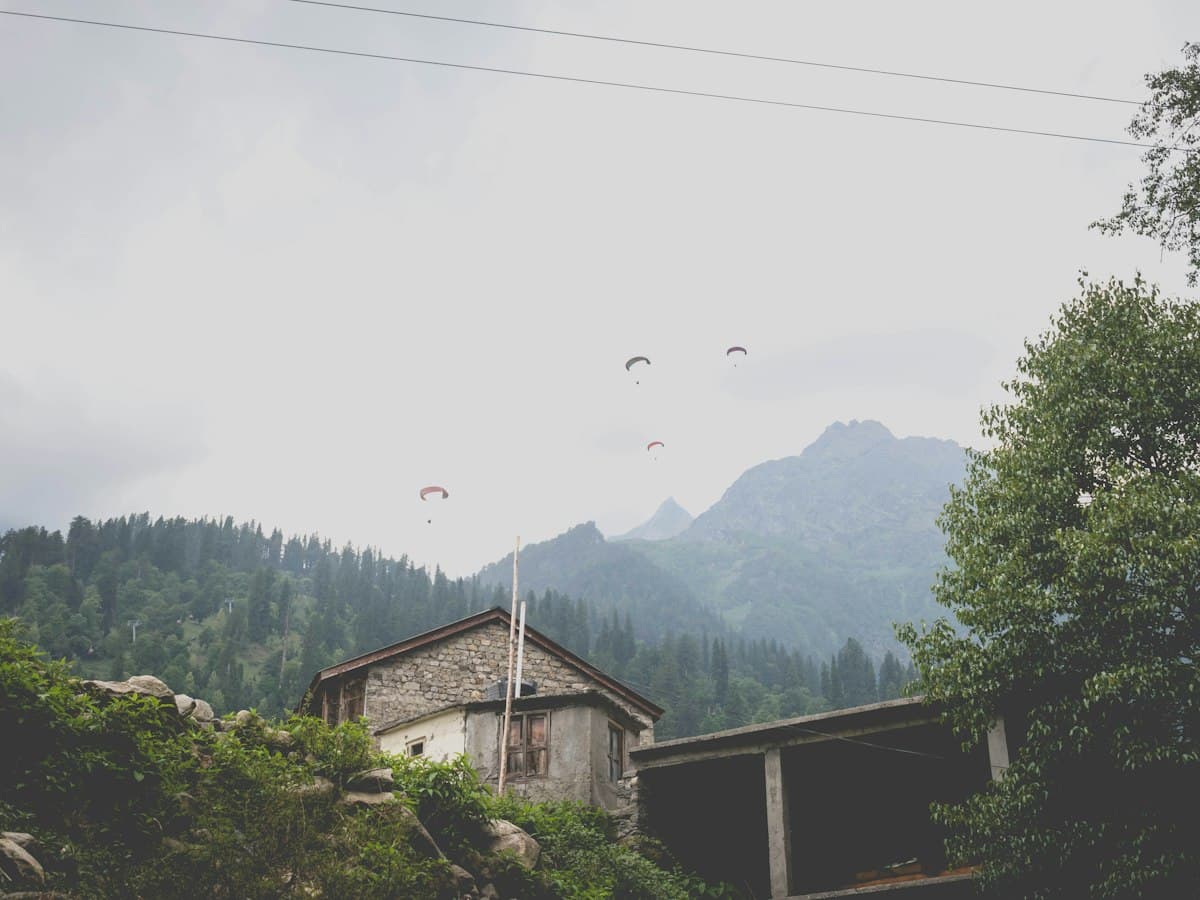 Several paragliders drift above a house in the Solang Valley near Manali with mountains around.