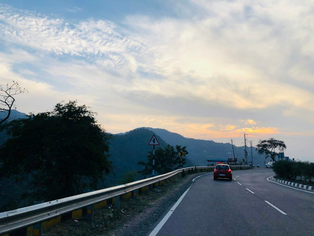 A car drives along a hill road with forested ridges and mountains in the background.