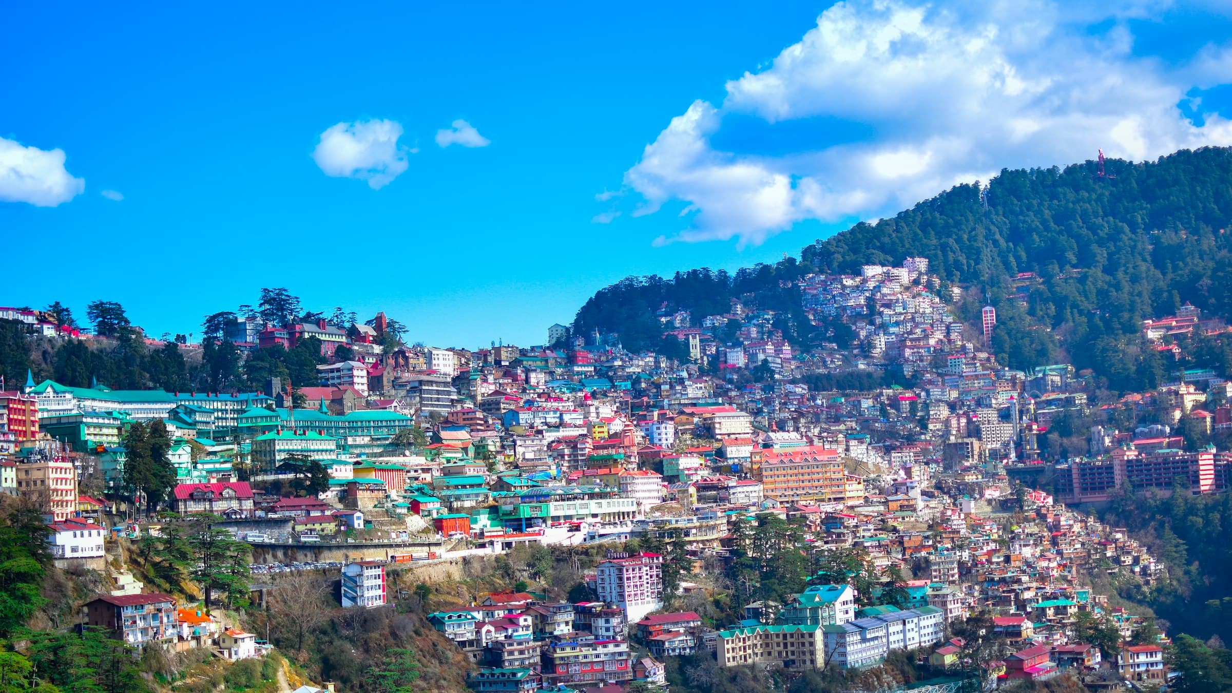 Shimla's hill-station skyline of stacked buildings sits below a mountain under a blue sky.