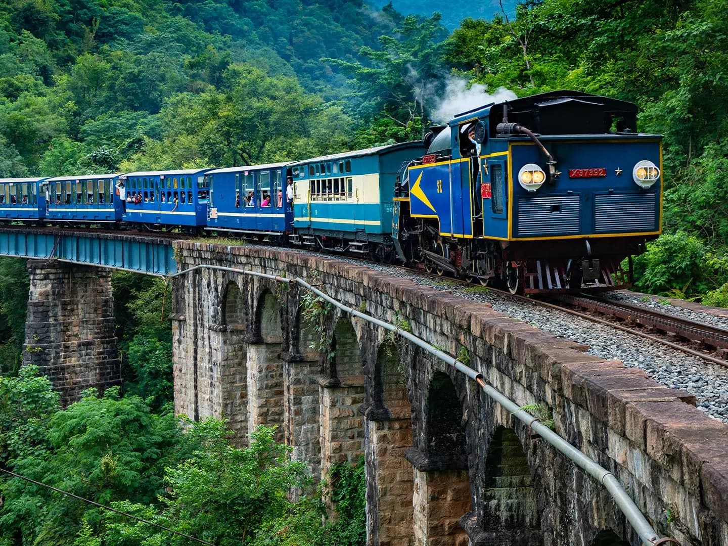 Passengers ride the blue-and-cream Nilgiri toy train as its steam engine crosses a stone-arch viaduct through green Nilgiri forest.