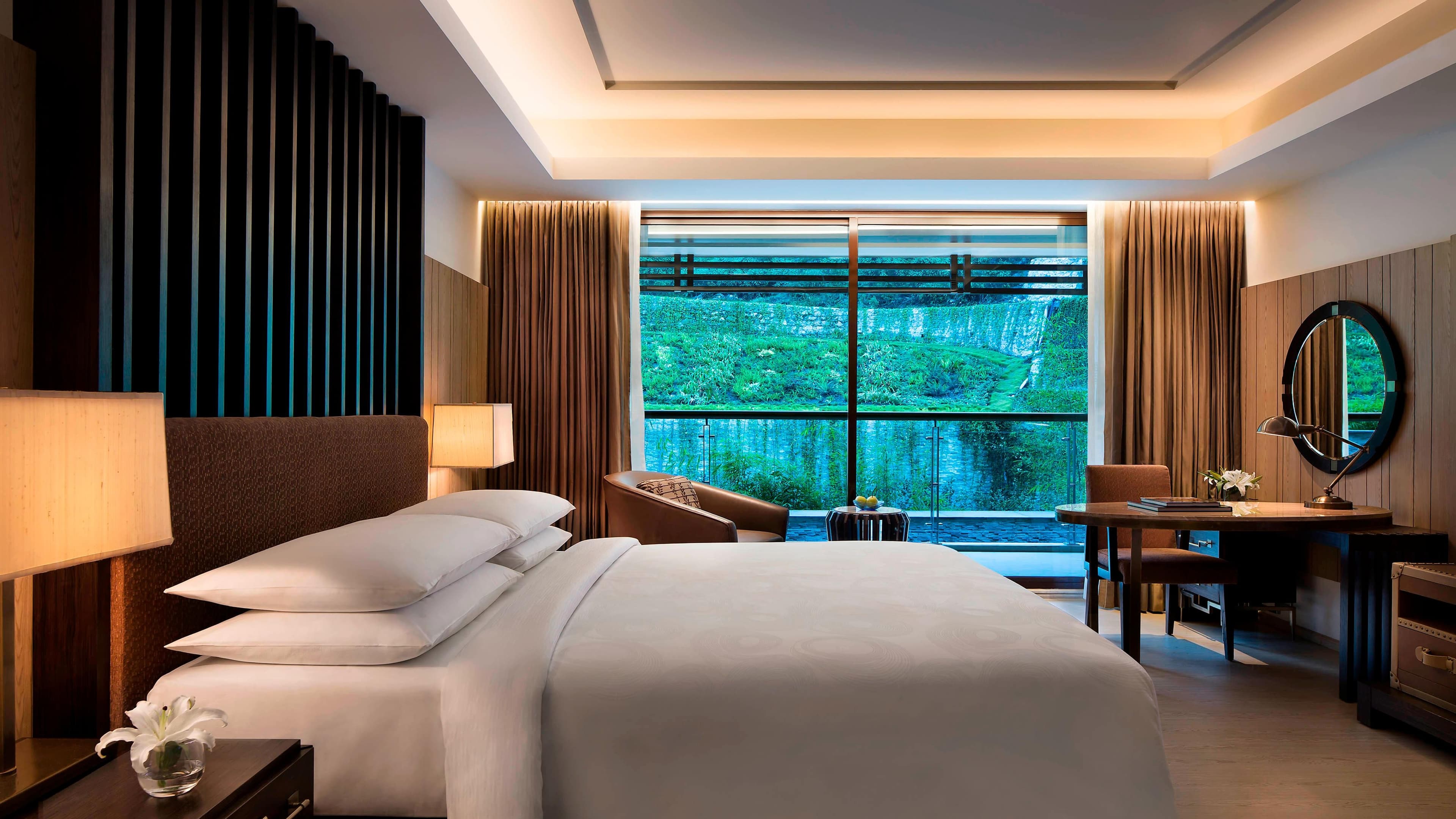 Guest room with king bed and private balcony overlooking a forested waterfall hillside at JW Marriott Mussoorie Walnut Grove Resort & Spa.