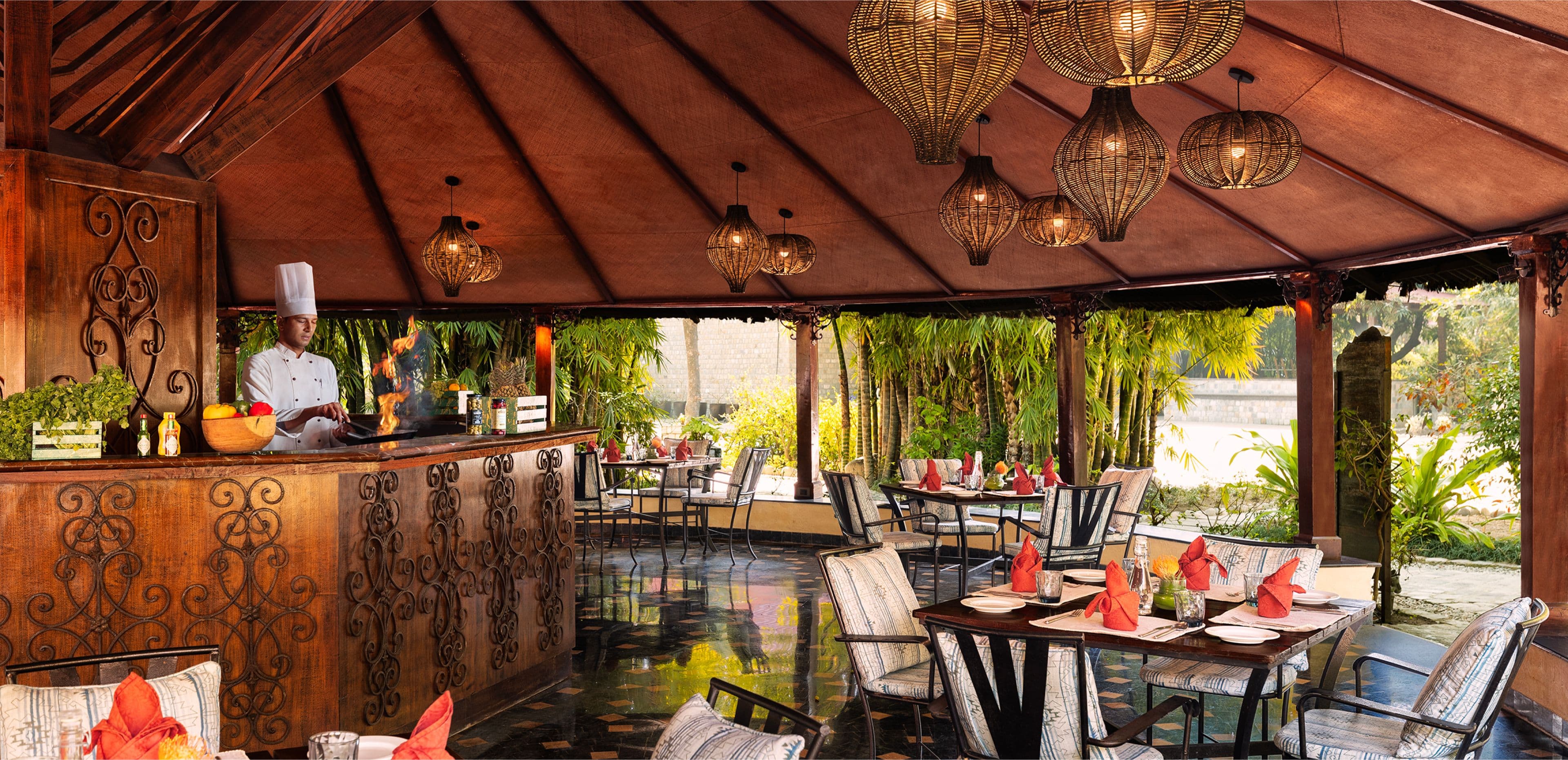 Open-air riverside restaurant pavilion at Taj Corbett Resort & Spa with a chef cooking at a live counter and rattan pendant lights