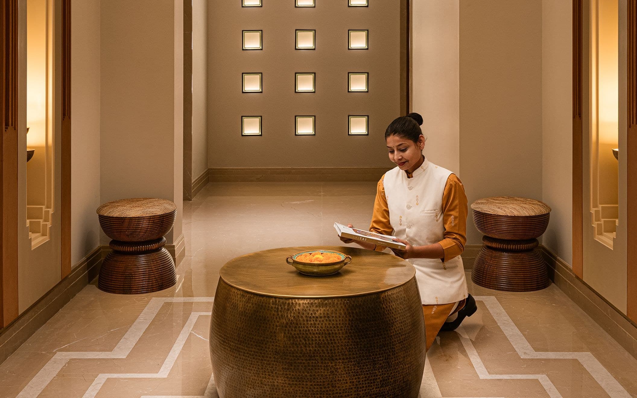 Haveli Hari Ganga spa corridor with a staff member in traditional attire carrying a welcome tray past illuminated wall niches