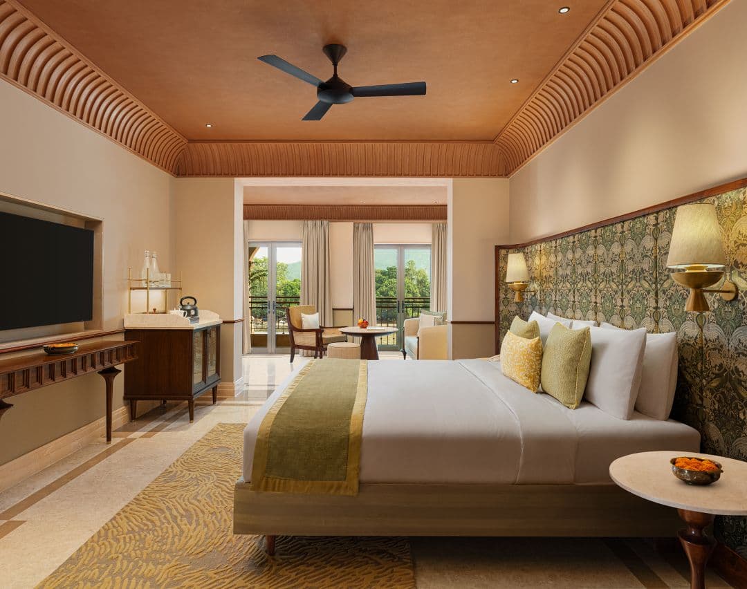 Haveli Hari Ganga guest room with king bed, patterned green headboard, wall-mounted TV, and a balcony overlooking greenery