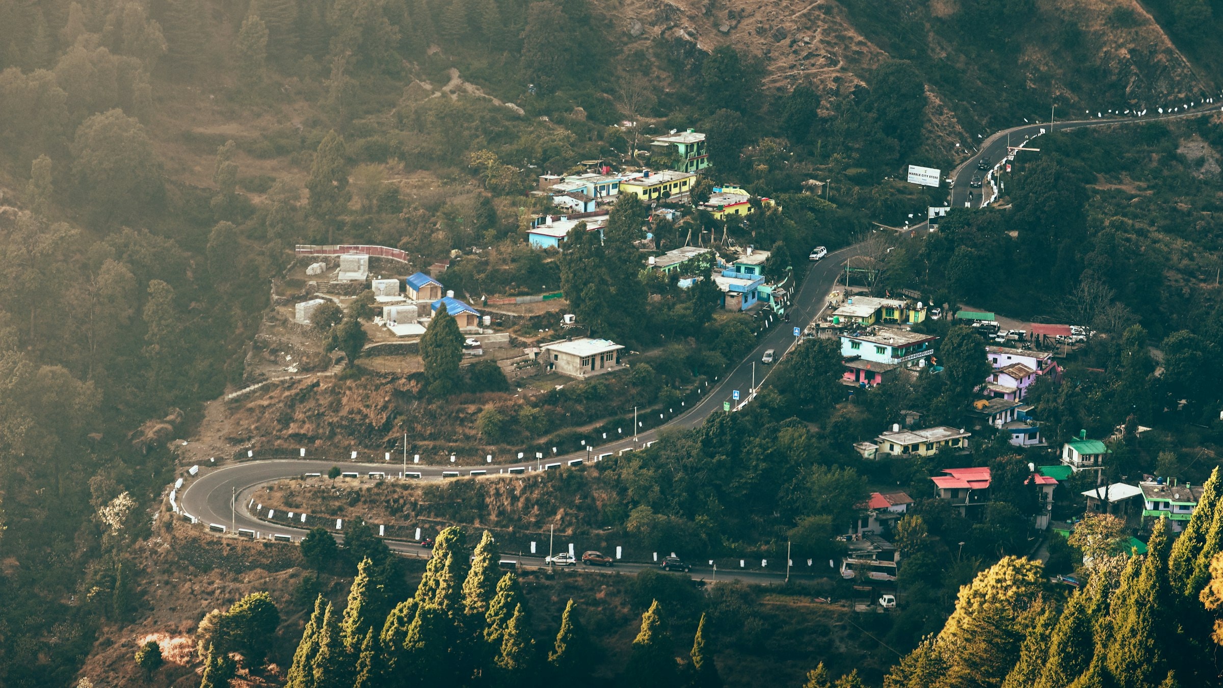 An aerial view of a winding mountain road and a hillside village surrounded by green hills and a valley beyond.