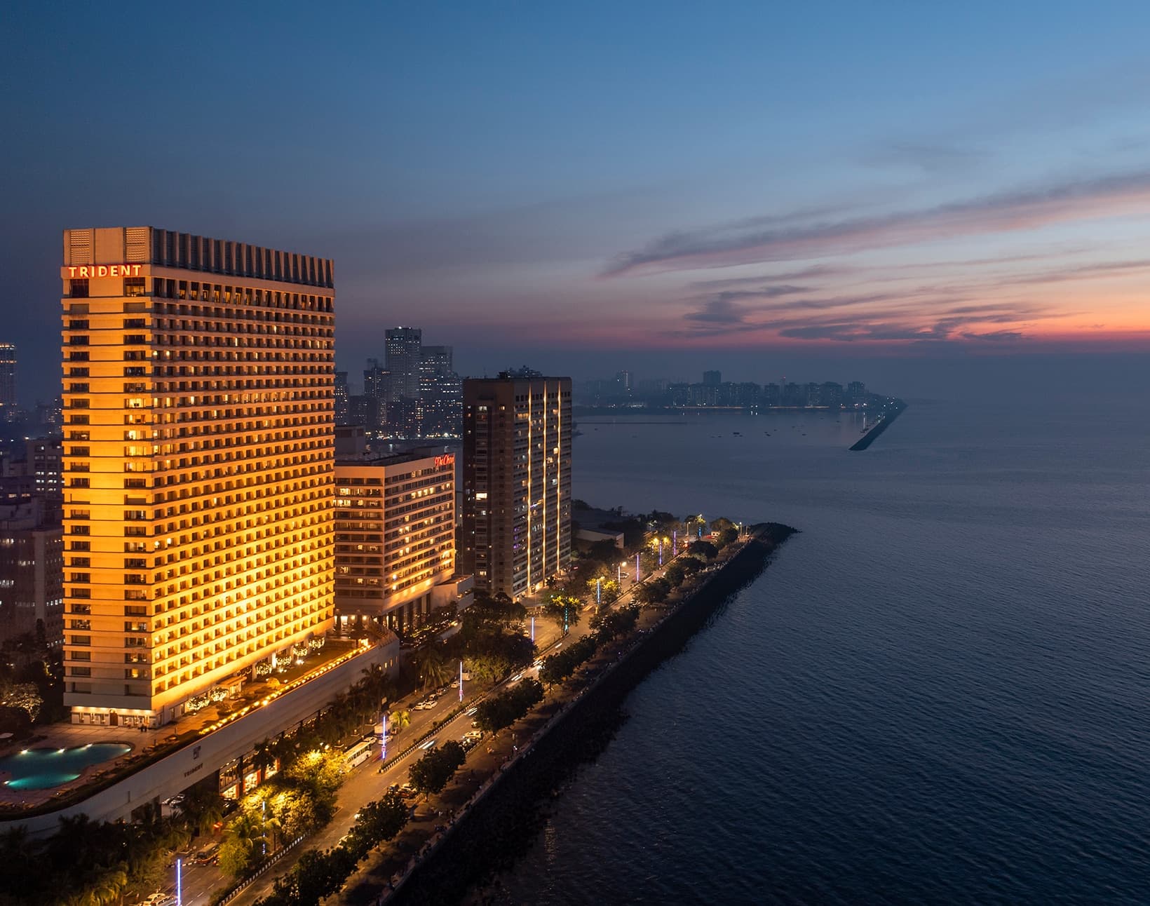 Trident, Nariman Point tower lit at dusk above the Marine Drive curve and the Arabian Sea